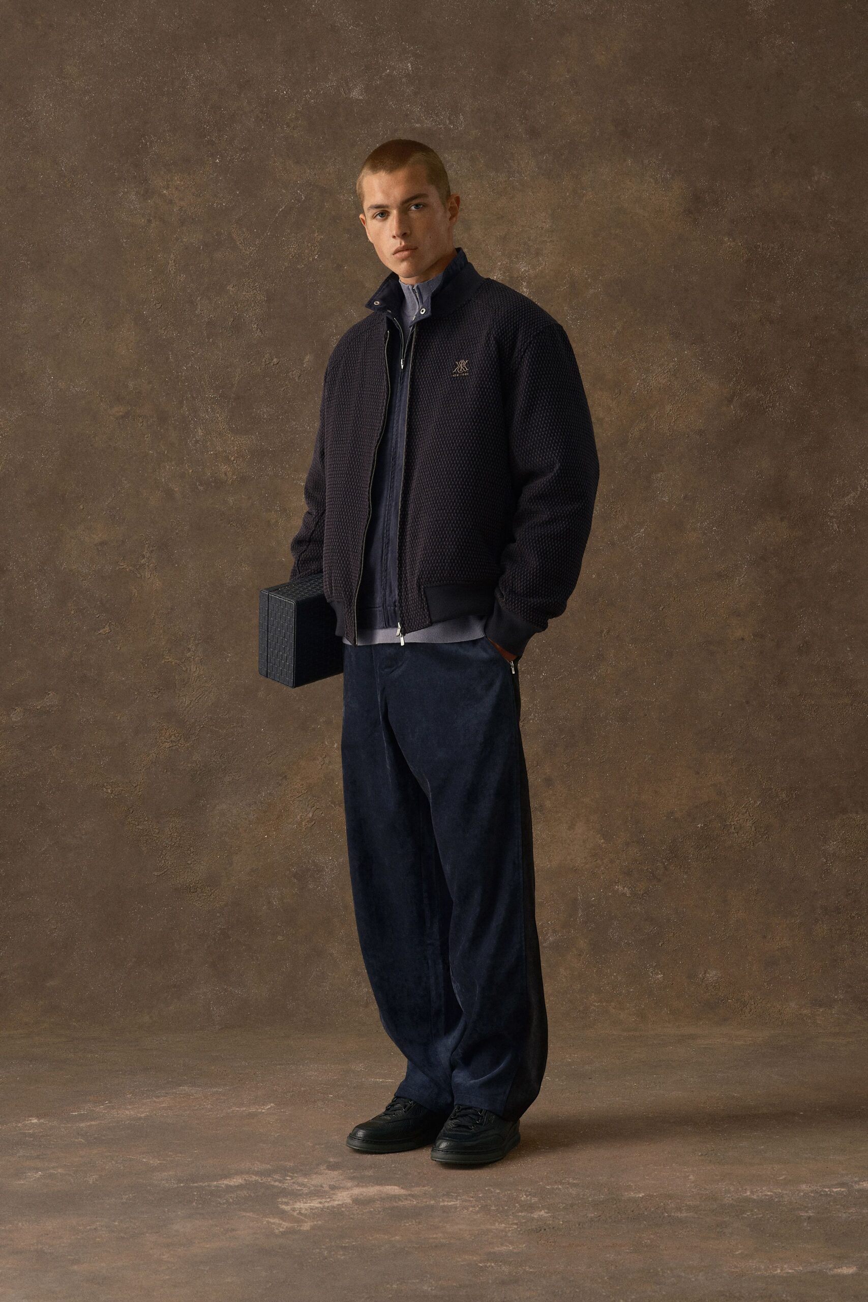 Navy bomber in a waffle-knit texture with ribbed cuffs, over a slate zip jacket and pale blue stand-collar shirt. Ink velvet trousers break over black leather sneakers; a boxy woven case in hand. Hotel bar, waiting on someone late.