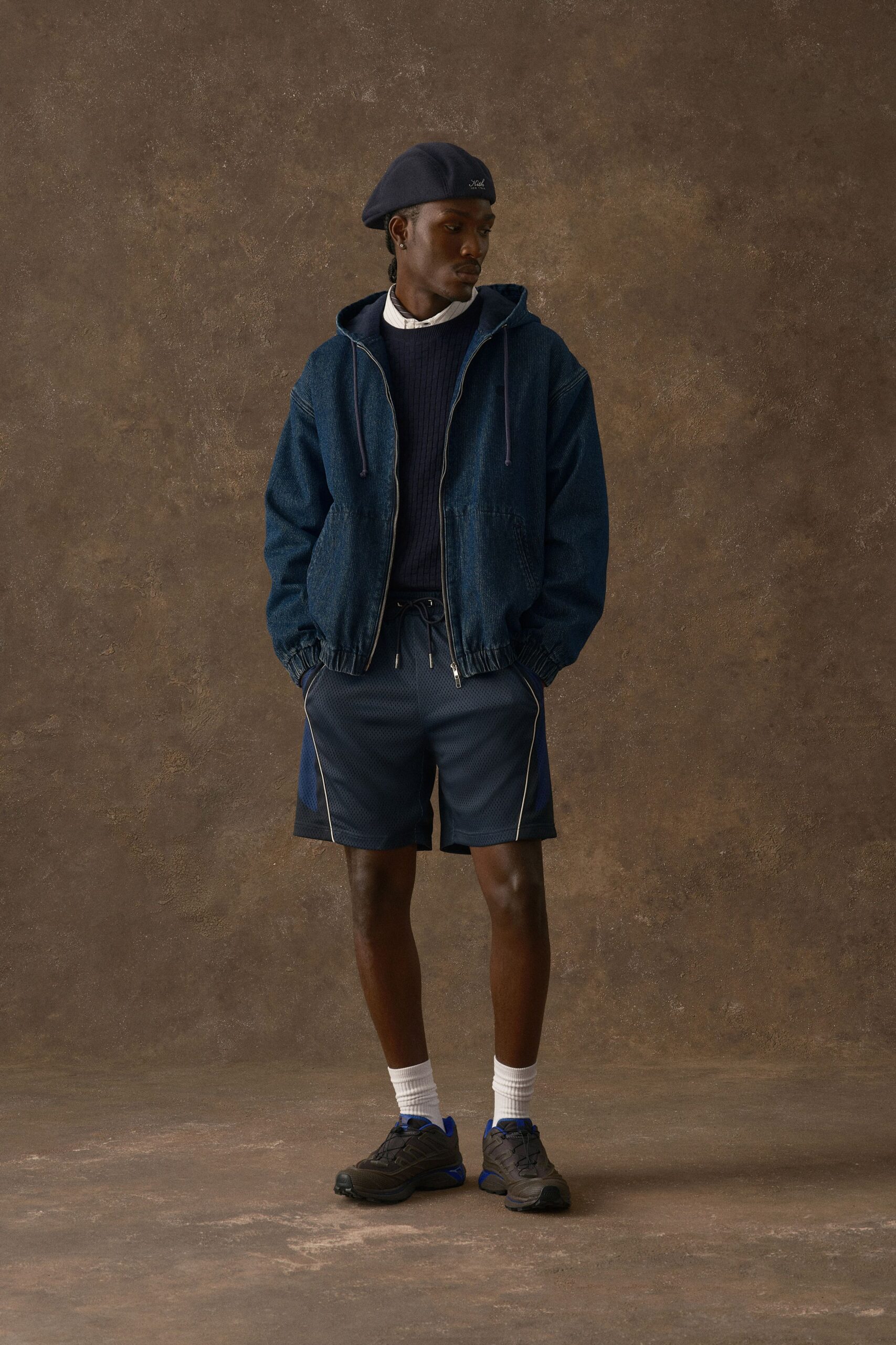 Navy wool beret, indigo denim zip hoodie with gathered cuffs, dark rib-knit sweater over a white band-collar shirt, mesh track shorts traced with pale piping, thick sport socks and trail sneakers. Waiting for the corner shop to open at dusk.