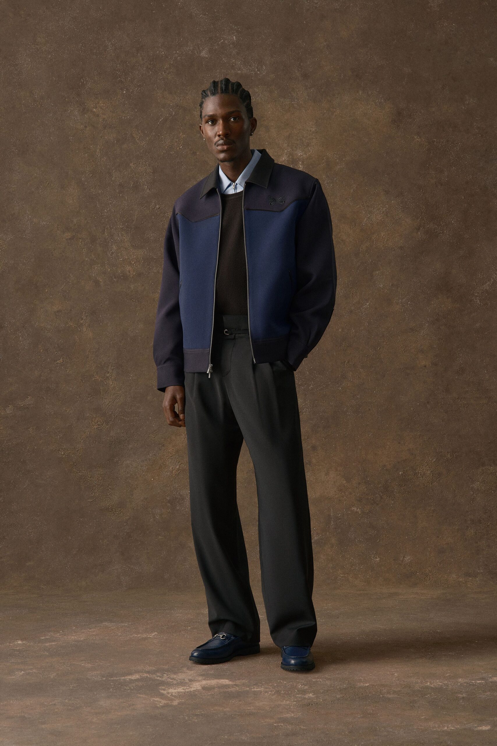 Navy zip-front jacket with a black yoke seam curving across the chest, over a pale blue poplin shirt and black crewneck knit. Charcoal pleated trousers with a side-tab waist, blue leather loafers. Waiting in a hotel bar before noon check-out.