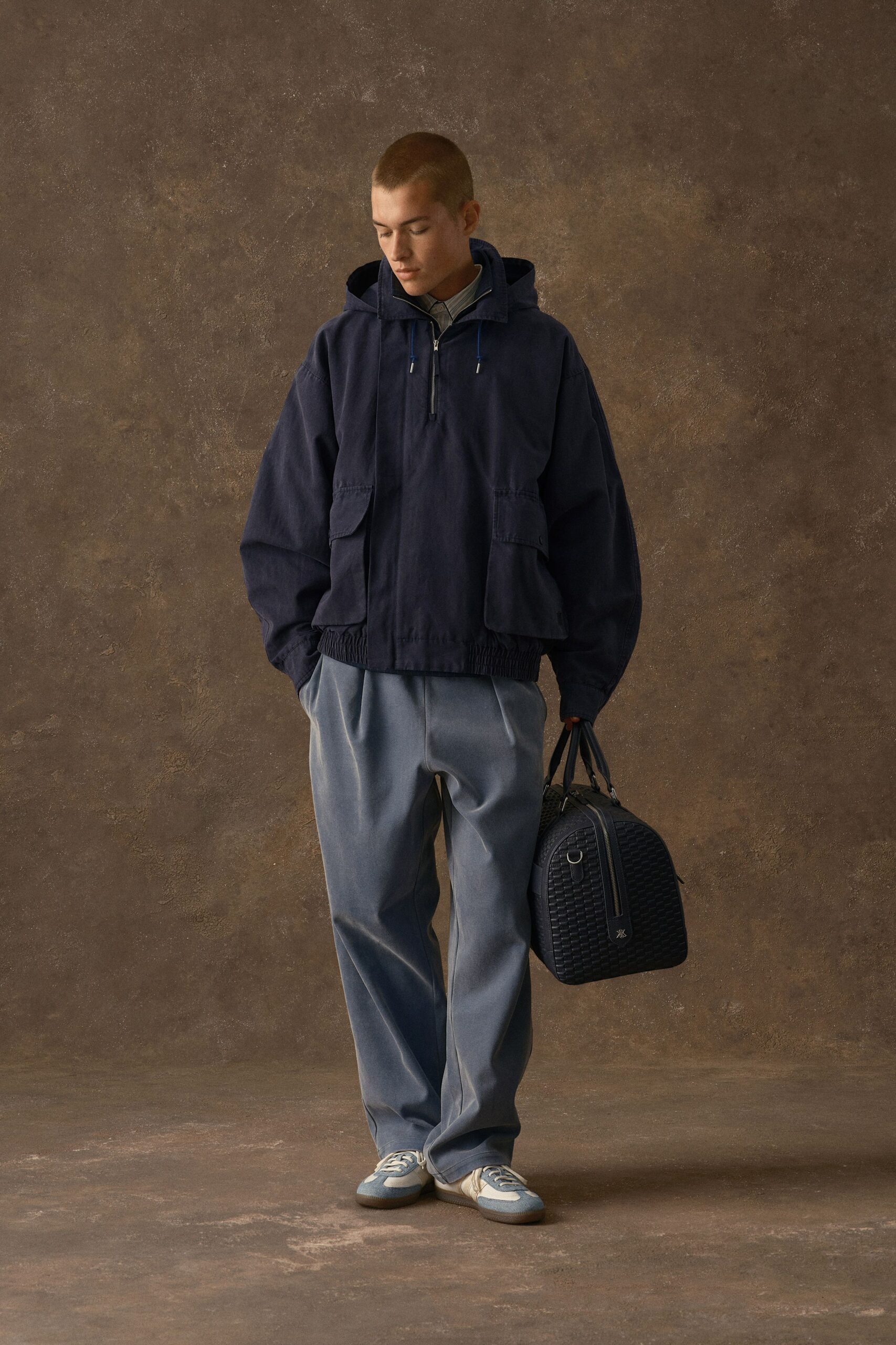 Navy hooded anorak in washed twill with a half zip and oversized flap pockets, pale blue stand-collar shirt beneath, slate trousers with a soft pressed crease, powder-blue trainers, woven leather holdall. Early train, platform tiles still damp.