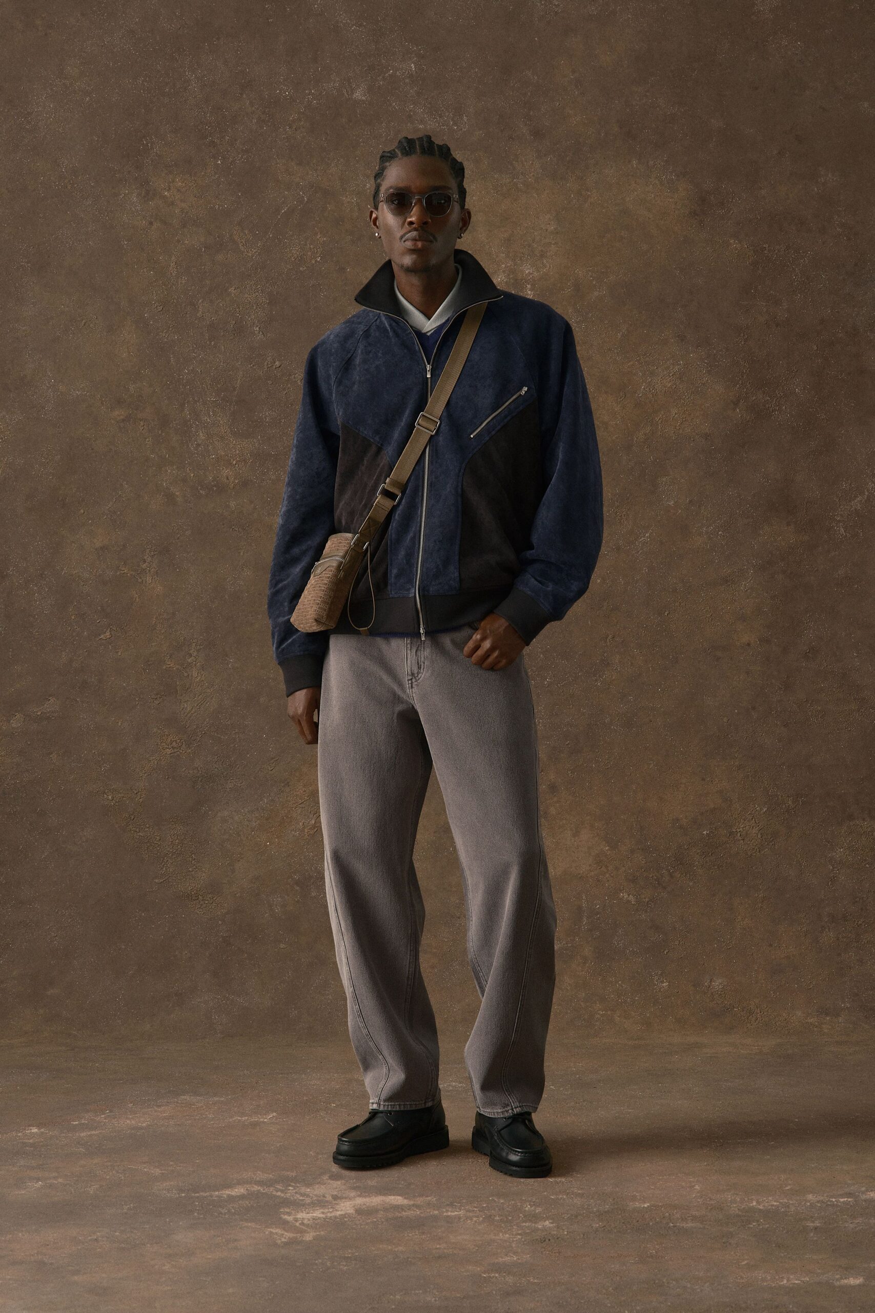 Indigo suede zip jacket with black contrast panels and a slanted chest zip, white poplin collar peeking over a cobalt V-neck, ash-wash jeans, black apron-toe loafers, tan embossed crossbody. Waiting in a hotel lobby that smells faintly of cedar.