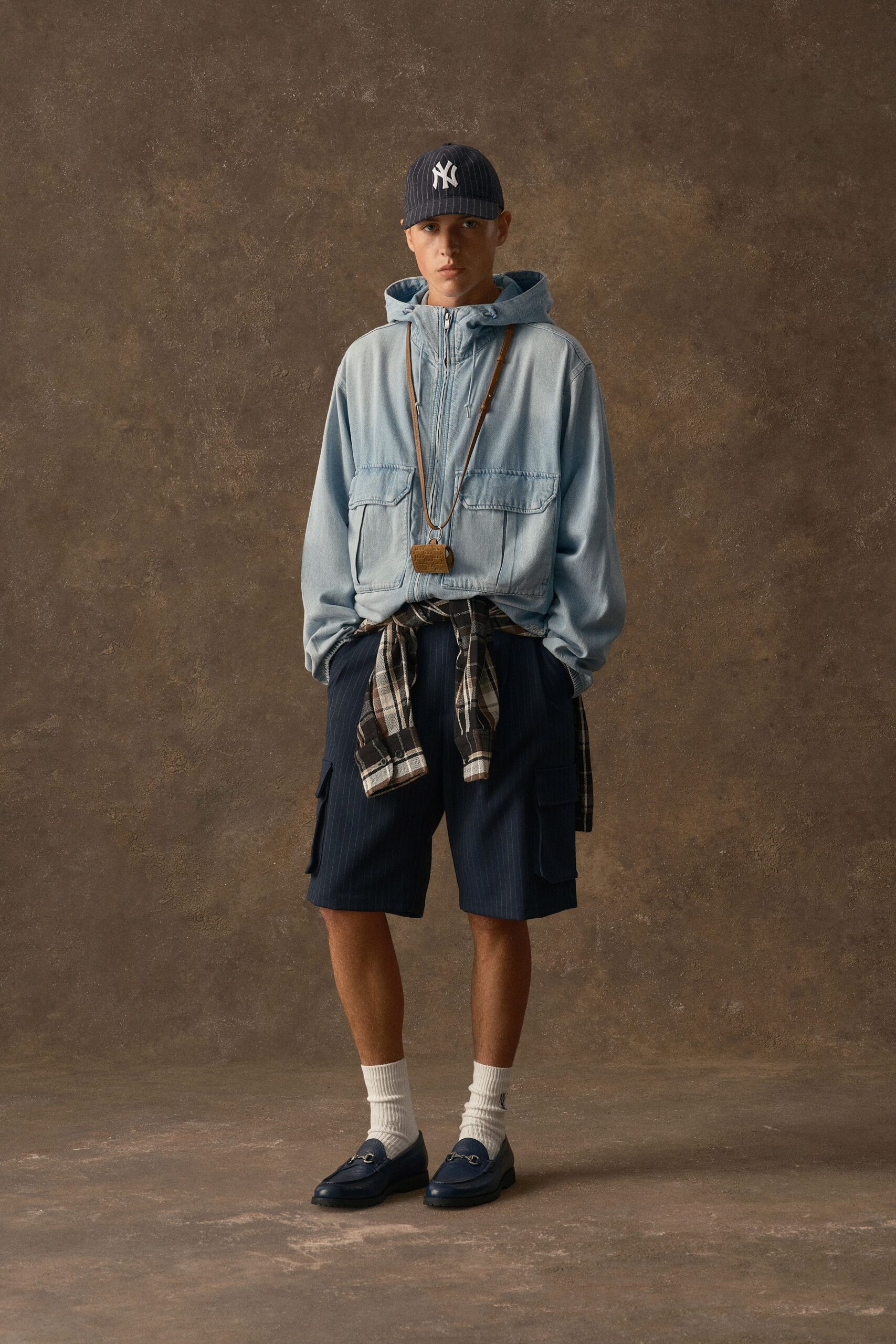 Navy pinstripe baseball cap, faded chambray hooded jacket with oversized flap pockets, plaid shirt knotted at the waist, navy pinstripe cargo shorts, ribbed white socks and horsebit loafers. Waiting outside a corner deli just after rain.