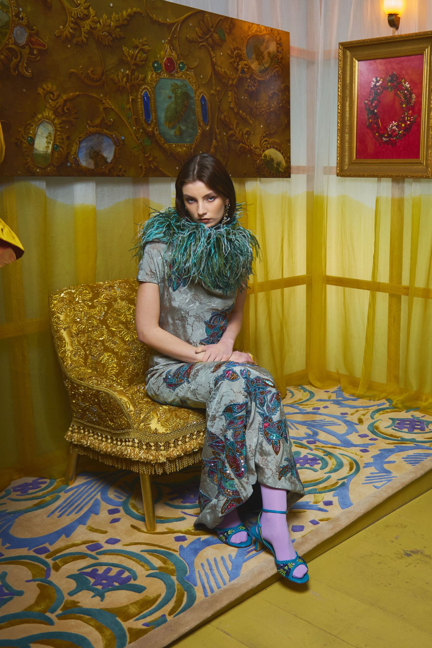 Silver jacquard column dress with short sleeves and beaded paisleys climbing the leg, ringed by a teal ostrich-feather collar. Lilac tights, turquoise ankle-strap sandals in matching beadwork. In a drawing room gone warm with lamp light.
