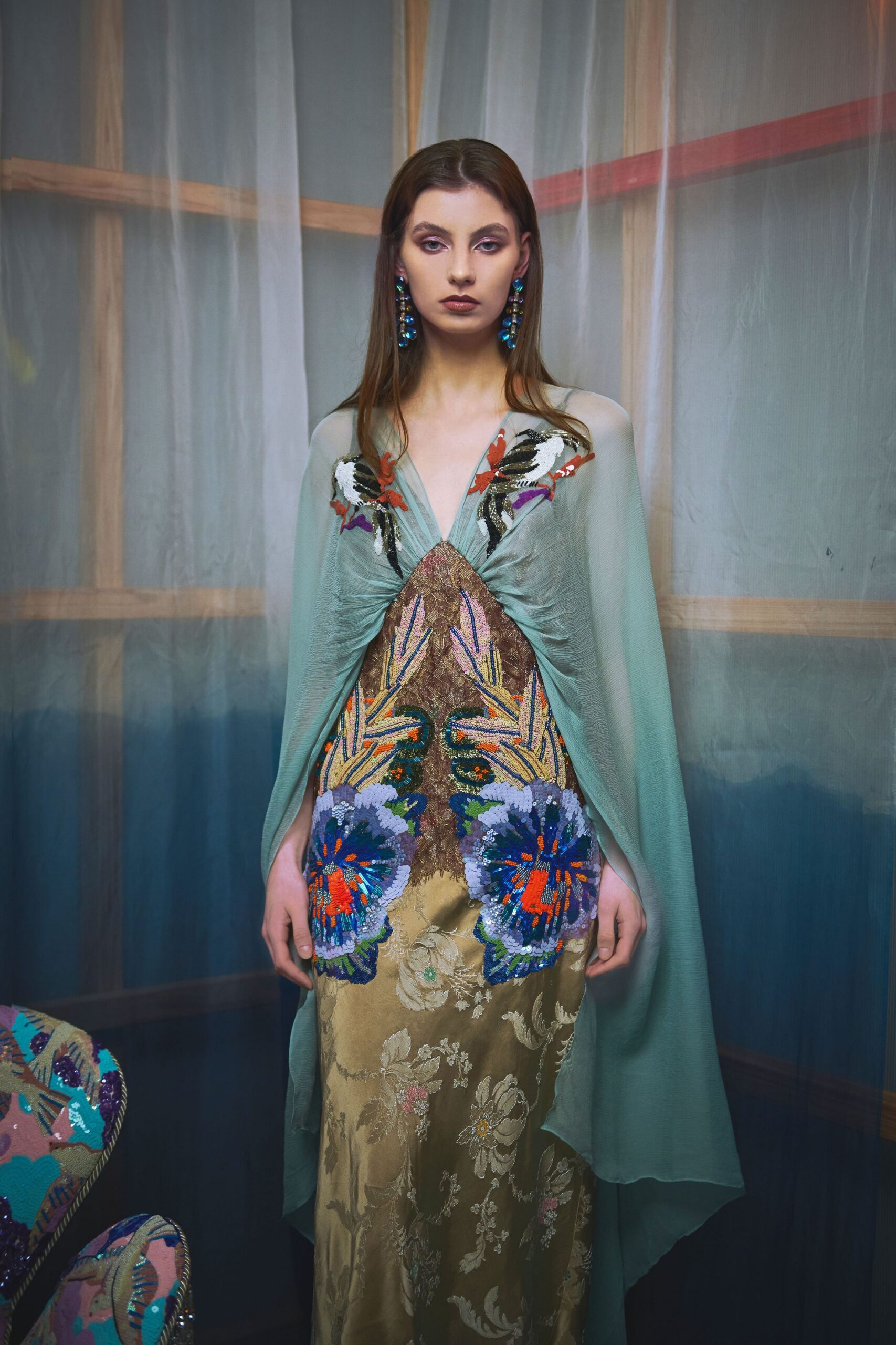 Seafoam chiffon cape dress, ruched into a deep V and scattered with sequin birds at the shoulders. Gold brocade skirt panel with heavy beadwork blooming into blue flowers. Waiting behind the theatre scrim.