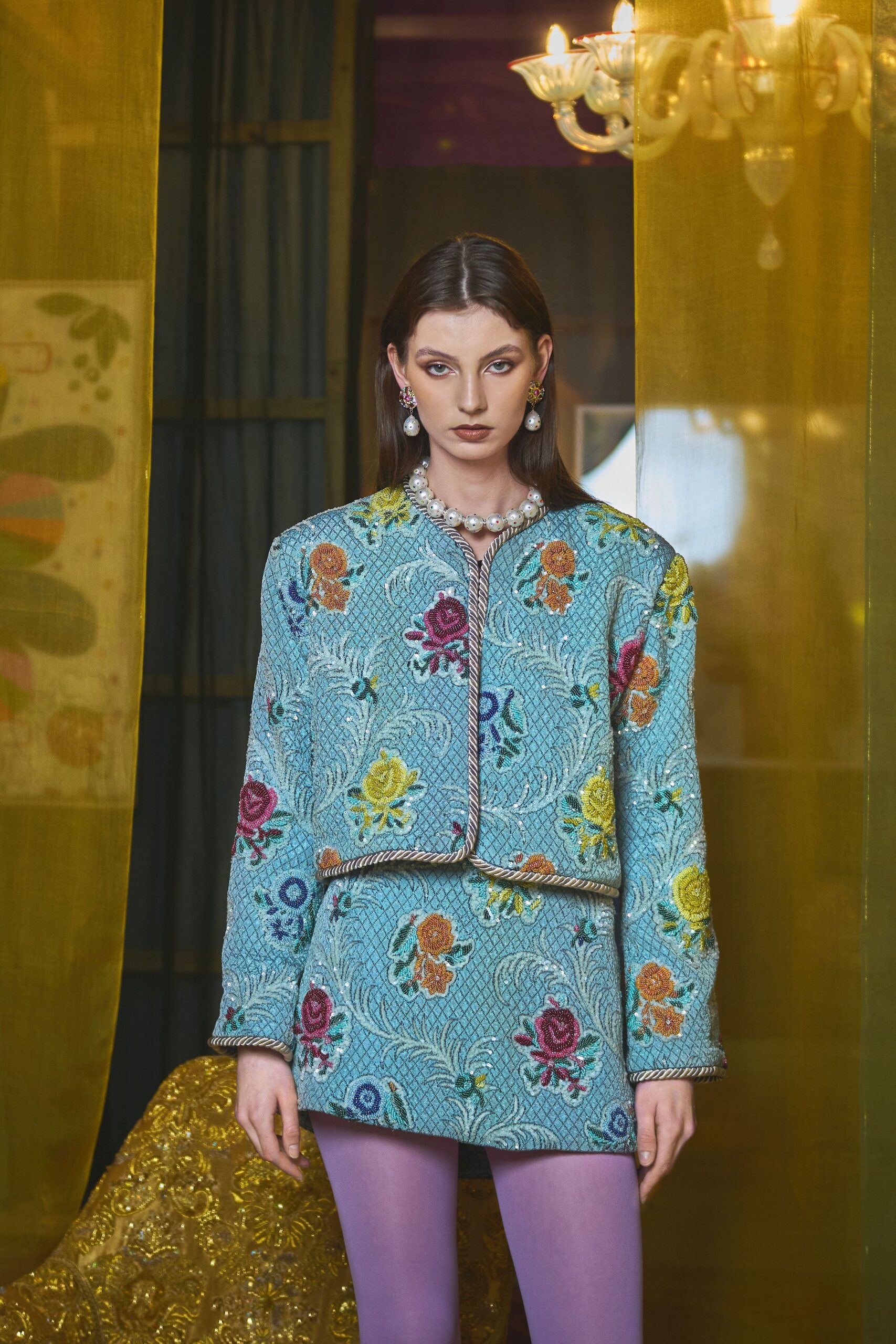 Powder-blue brocade jacket traced with twisted cord piping, over a matching mini skirt dense with raised floral embroidery. Lavender tights, baroque-pearl drop earrings, chunky pearl necklace. Waiting in a hotel salon after midnight.