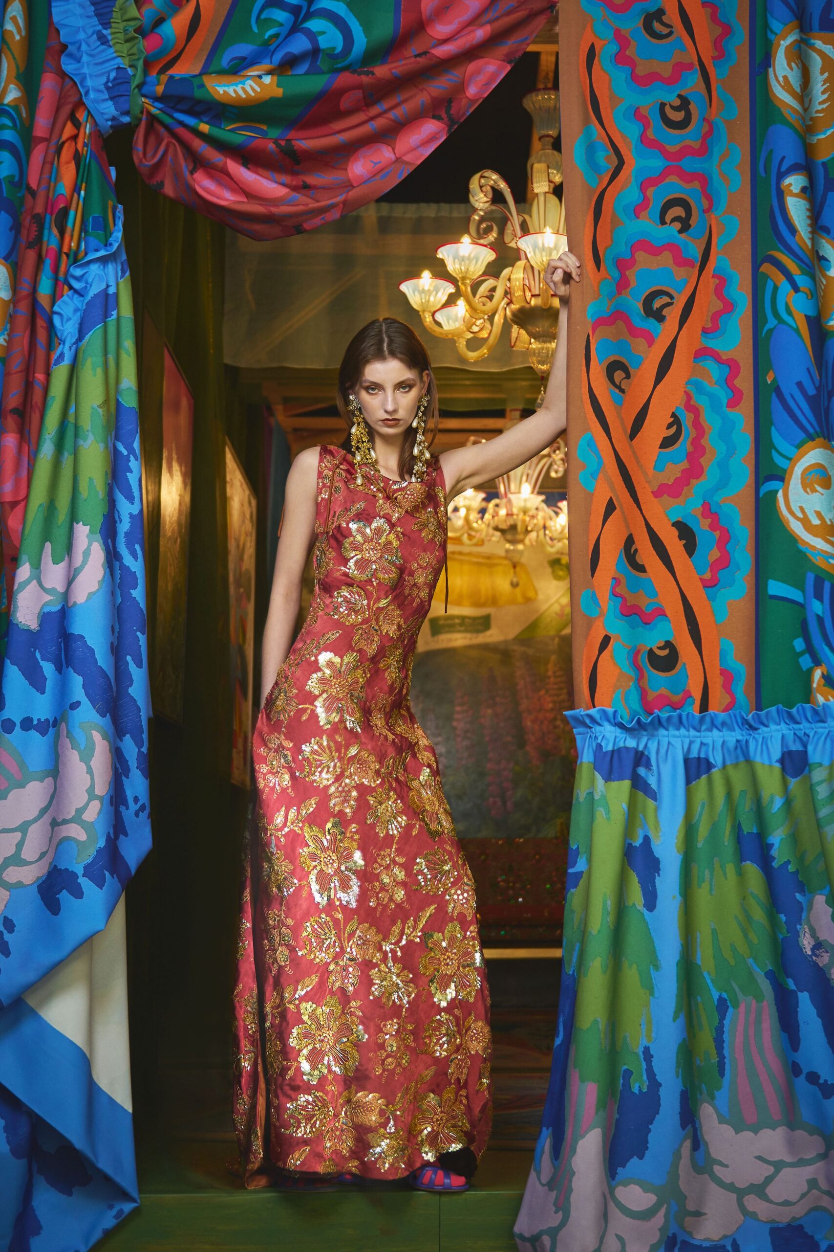 Sleeveless column gown in rose silk, dense with gold floral beading that catches at every curve. Long chandelier earrings with pearl drops. A look for slipping through a velvet-curtained hallway after midnight.