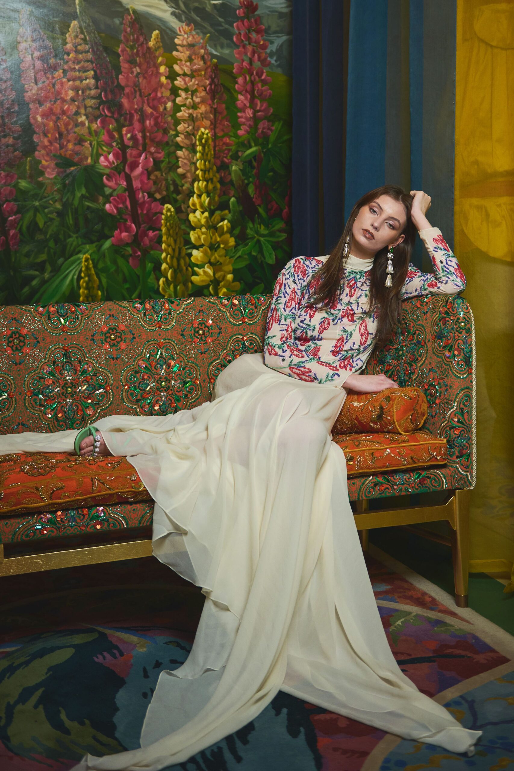 Ivory mock-neck top densely beaded with coral and cobalt sprigs, over an ivory georgette skirt that puddles into a long train. Mint sandals with crystal straps, tassel earrings. Waiting out dessert in a hotel drawing room.