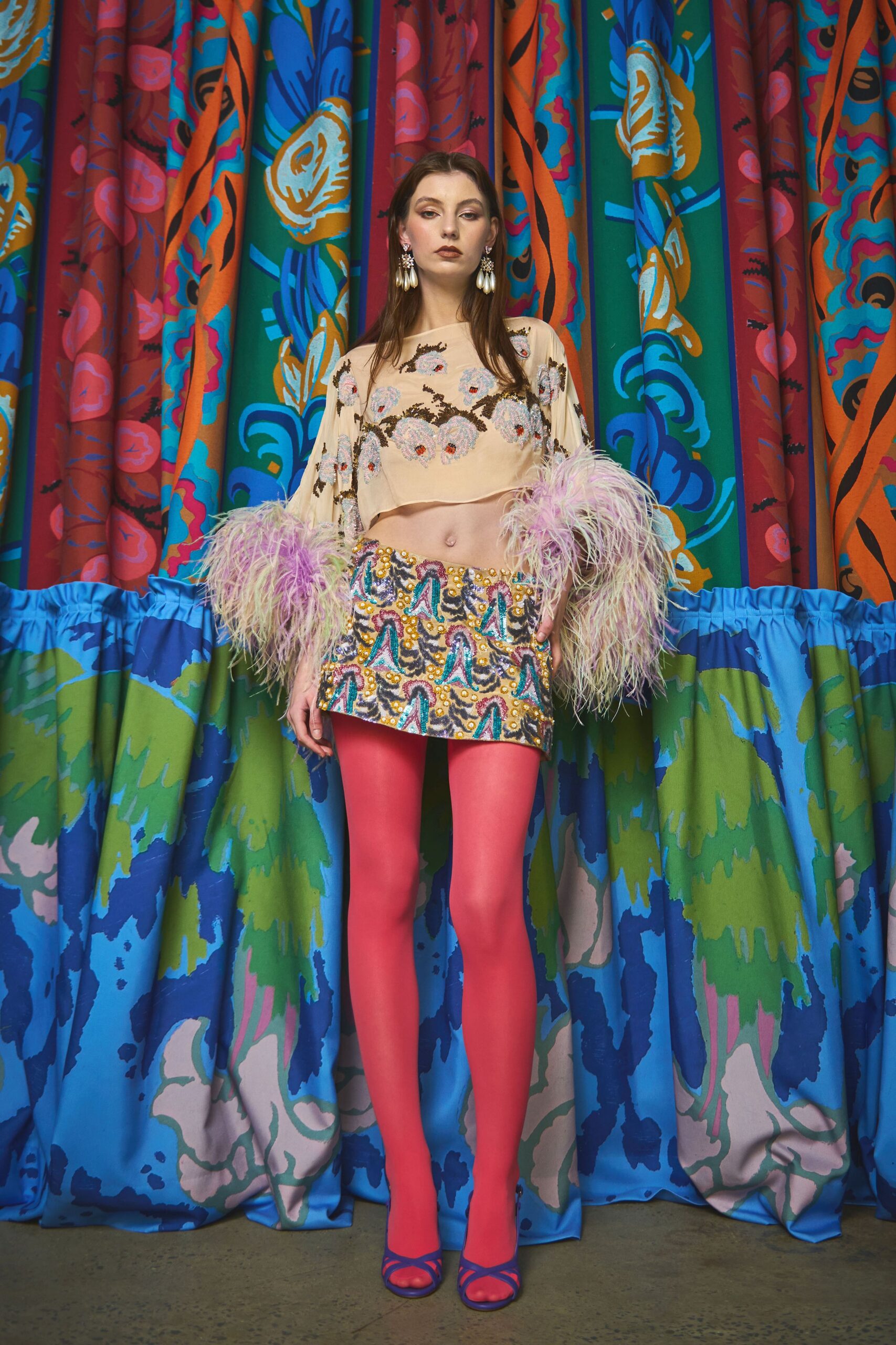 Cropped blush top with beaded blossom embroidery and ostrich-feather cuffs, low on the ribcage. Beaded mini skirt in a zigzag motif, hot-pink tights, cobalt strappy heels. Waiting by the velvet curtain after midnight karaoke.
