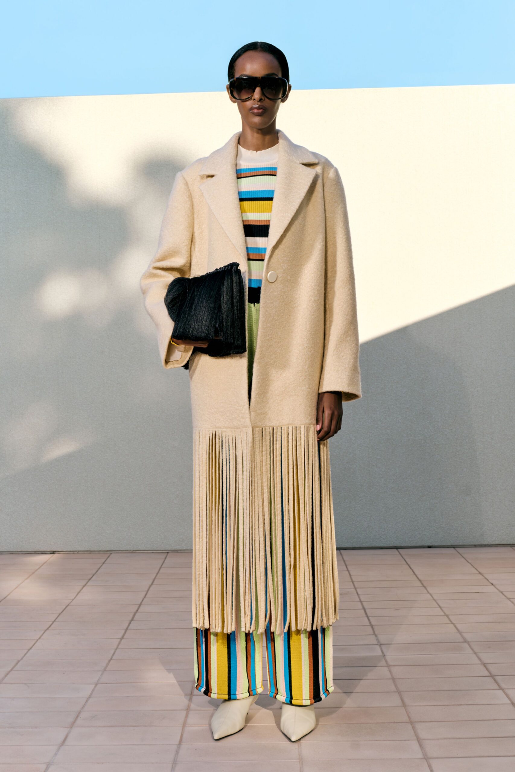 Cream brushed-wool coat with a curtain of long fringe at the hem, over a ribbed knit set striped in citron, sky and black. Black pleated clutch, ivory pointed boots, oversize gradient shades. Sun hitting a tiled courtyard after lunch.