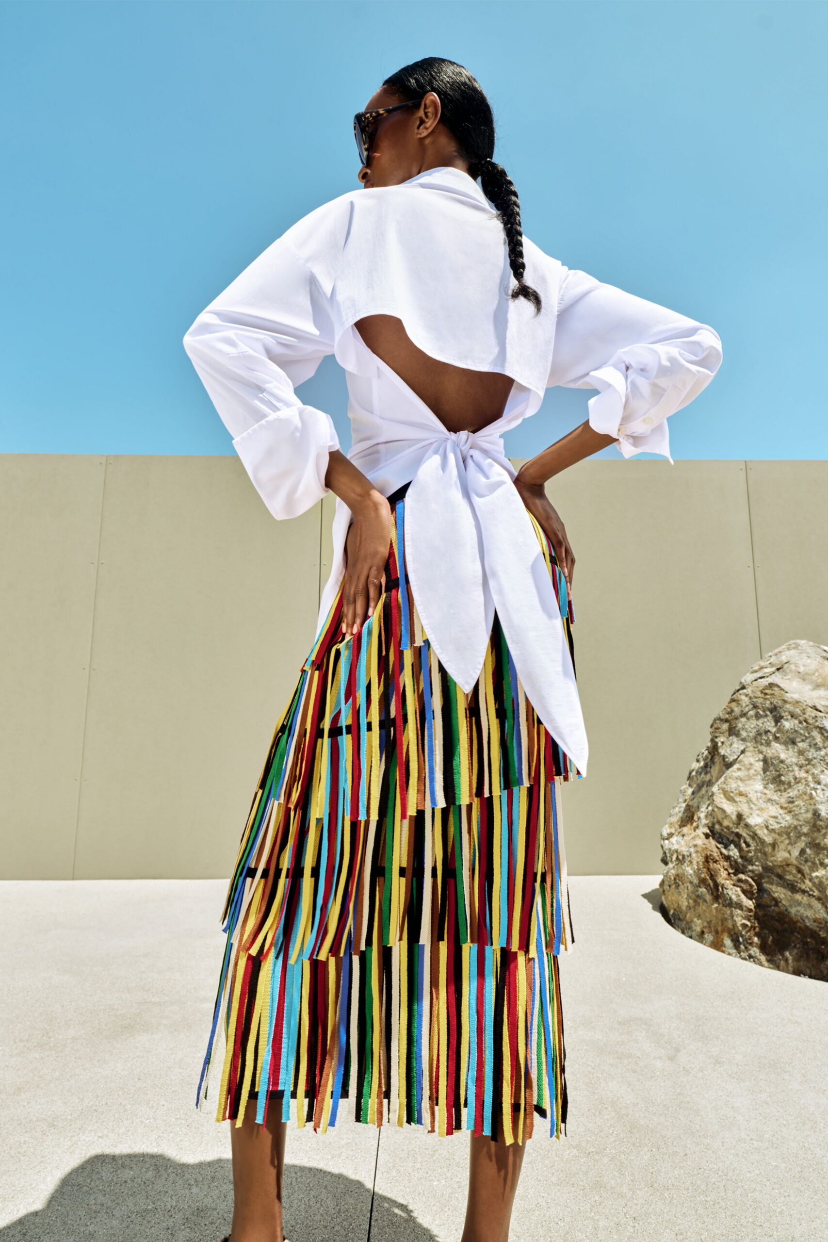 Tortoiseshell cat-eye sunglasses with thick glossy temples. White cotton blouse cut open across the back, tied low in a bow. Midi skirt covered in swinging grosgrain strips, layered in uneven tiers. Walking along a museum terrace at noon, wind lifting every ribbon end.