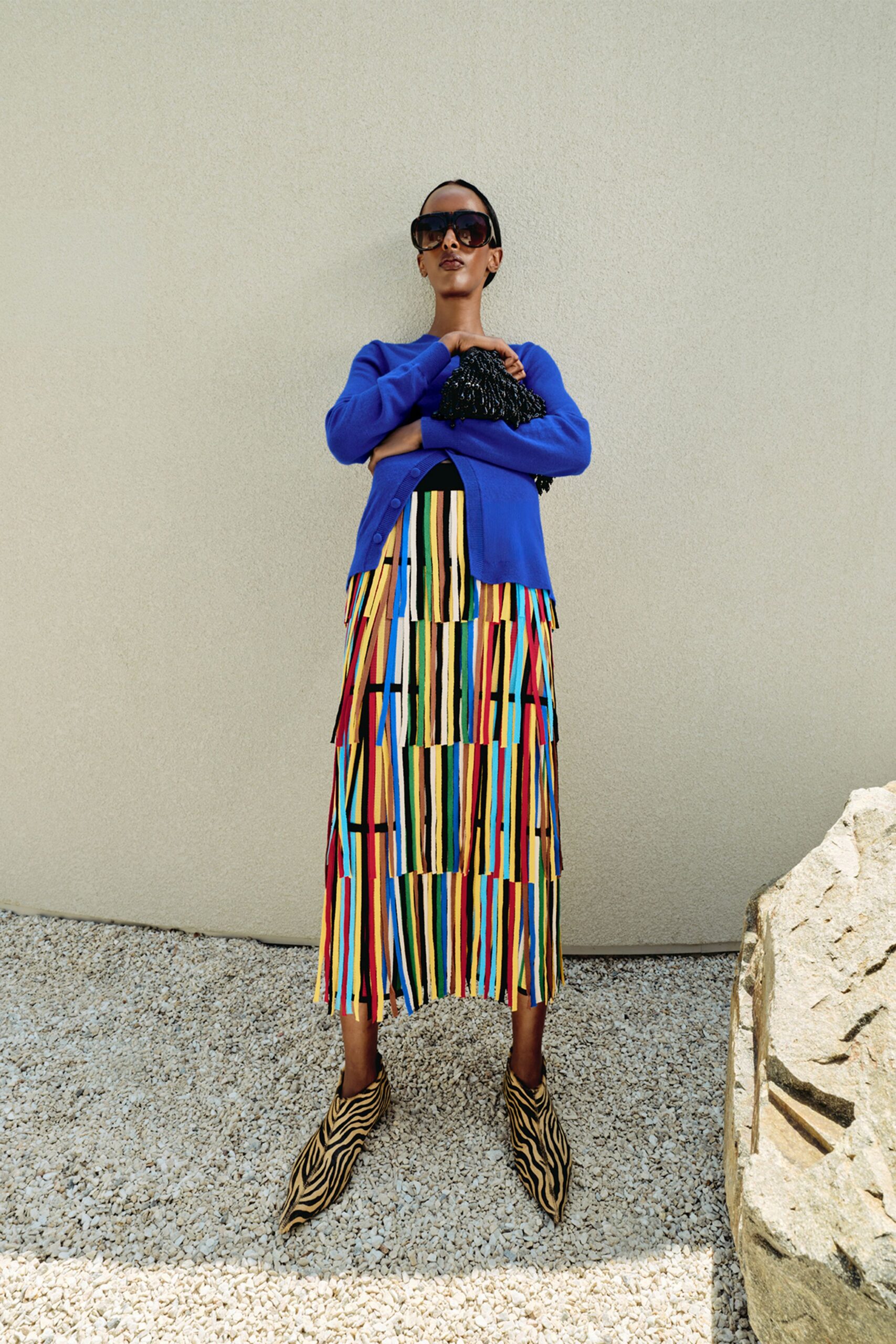 Cobalt knit cardigan with a neat row of covered buttons, over a midi skirt built from dangling multicolour ribbon strips. Black beaded pouch tucked at the chest, zebra-print pointed boots. Noon on a gravel courtyard by a sunstruck wall.