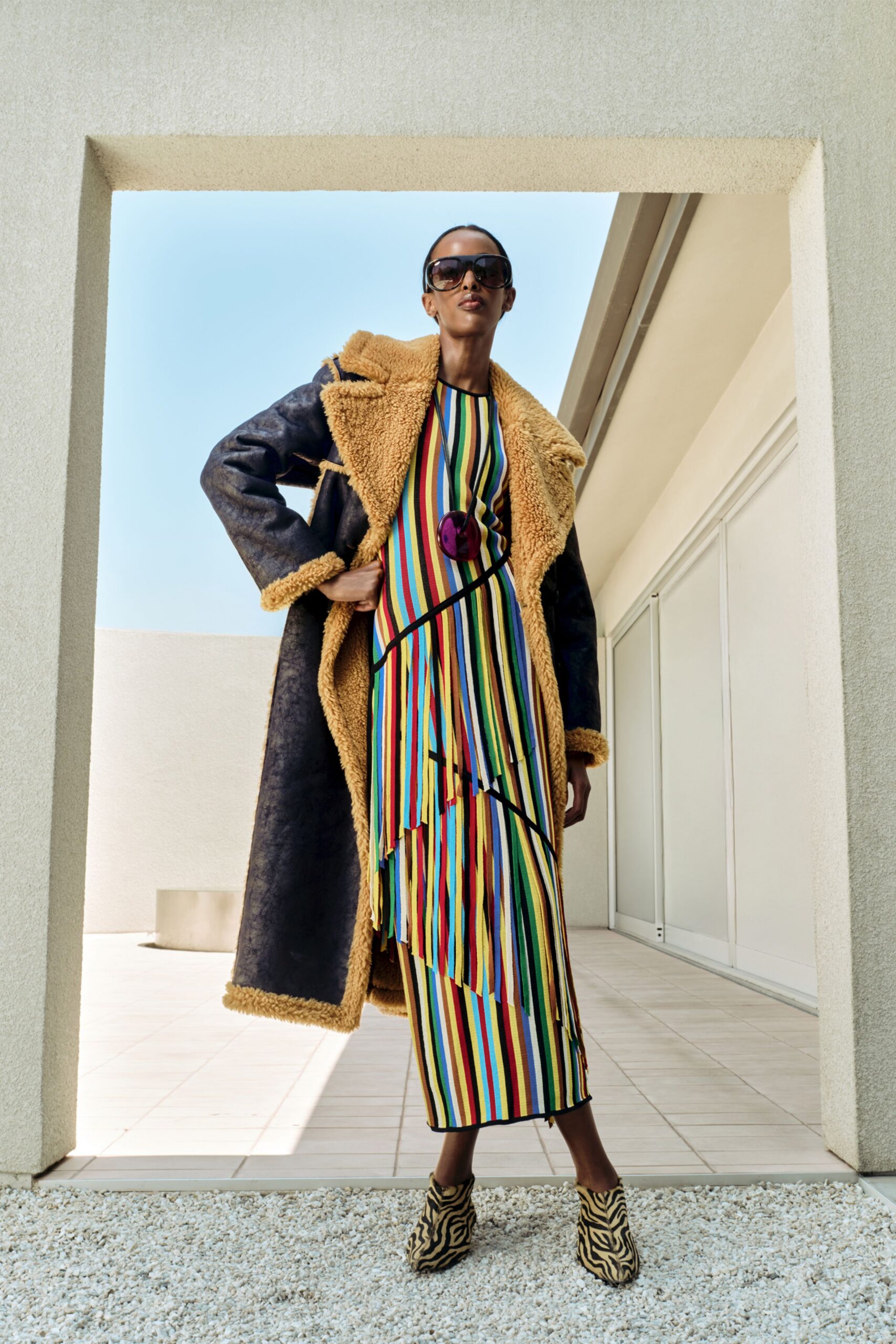 Long black shearling coat with a dense caramel pile at the lapels and cuffs. Ribbed knit dress in narrow candy-striped columns, cut with slanting fringe panels. Zebra calf-hair ankle boots. Sun on the tiles of a rented villa terrace.