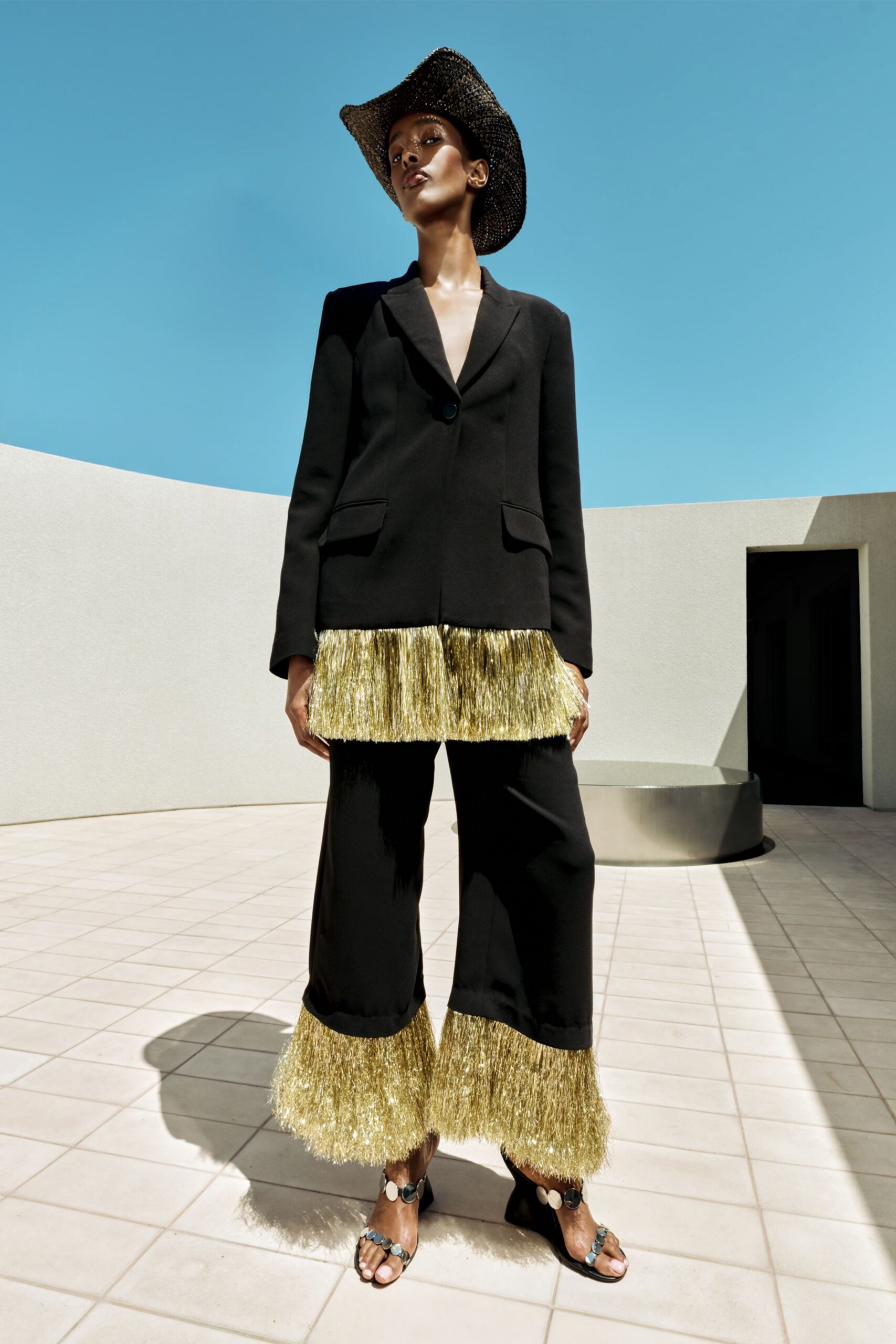 Black single-button blazer with a deep notch lapel, its hem finished in gold tinsel fringe. Cropped black trousers with matching tinsel at the cuff, black sandals studded with silver discs, woven cowboy hat. Noon by a drained courtyard fountain.