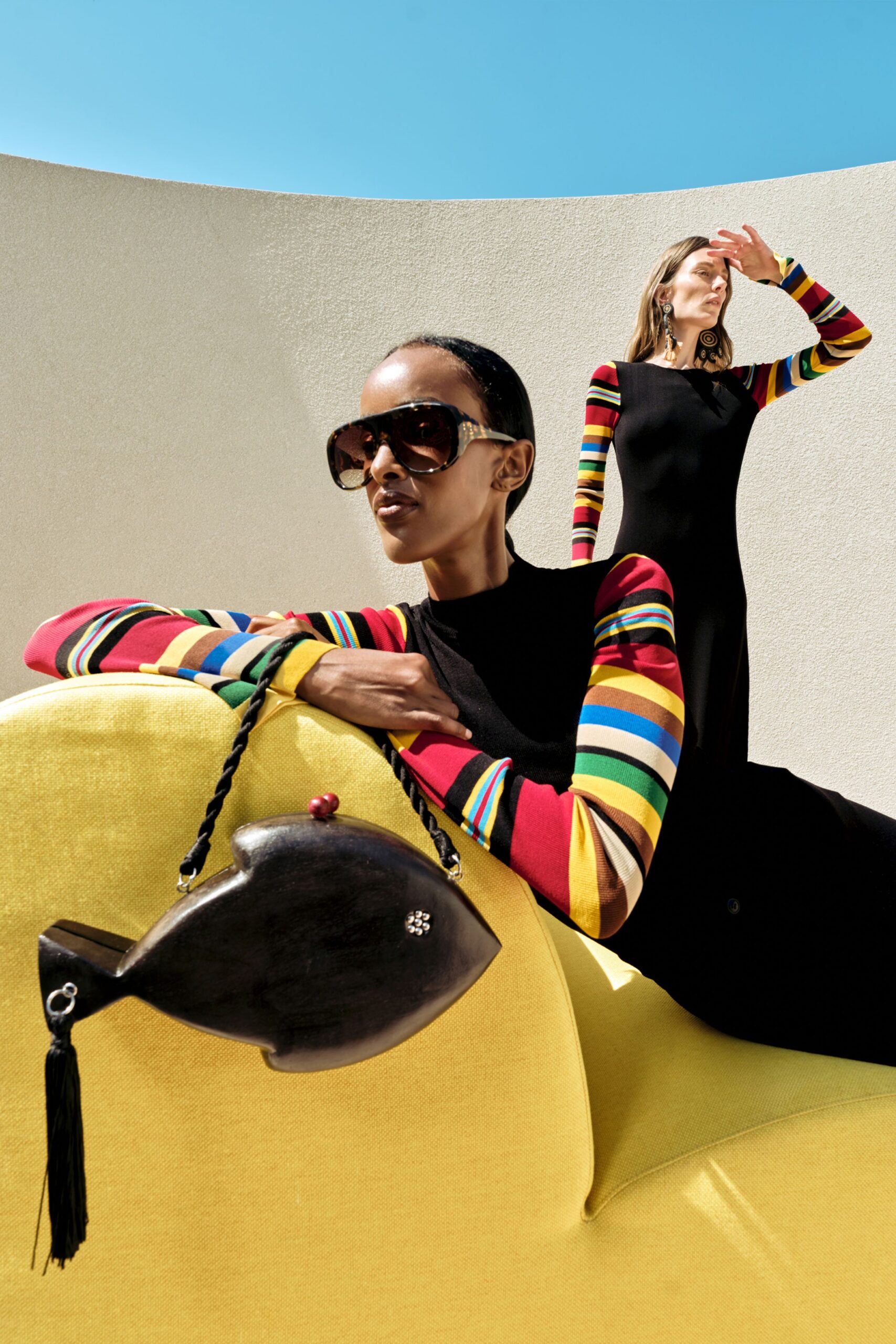 Black column dress in fine rib knit, its long sleeves banded in candy-bright stripes. Oversized tortoiseshell sunglasses, fish-shaped wooden bag with a twisted cord handle and tassel. Noon by the stucco pool wall, squinting into heat.