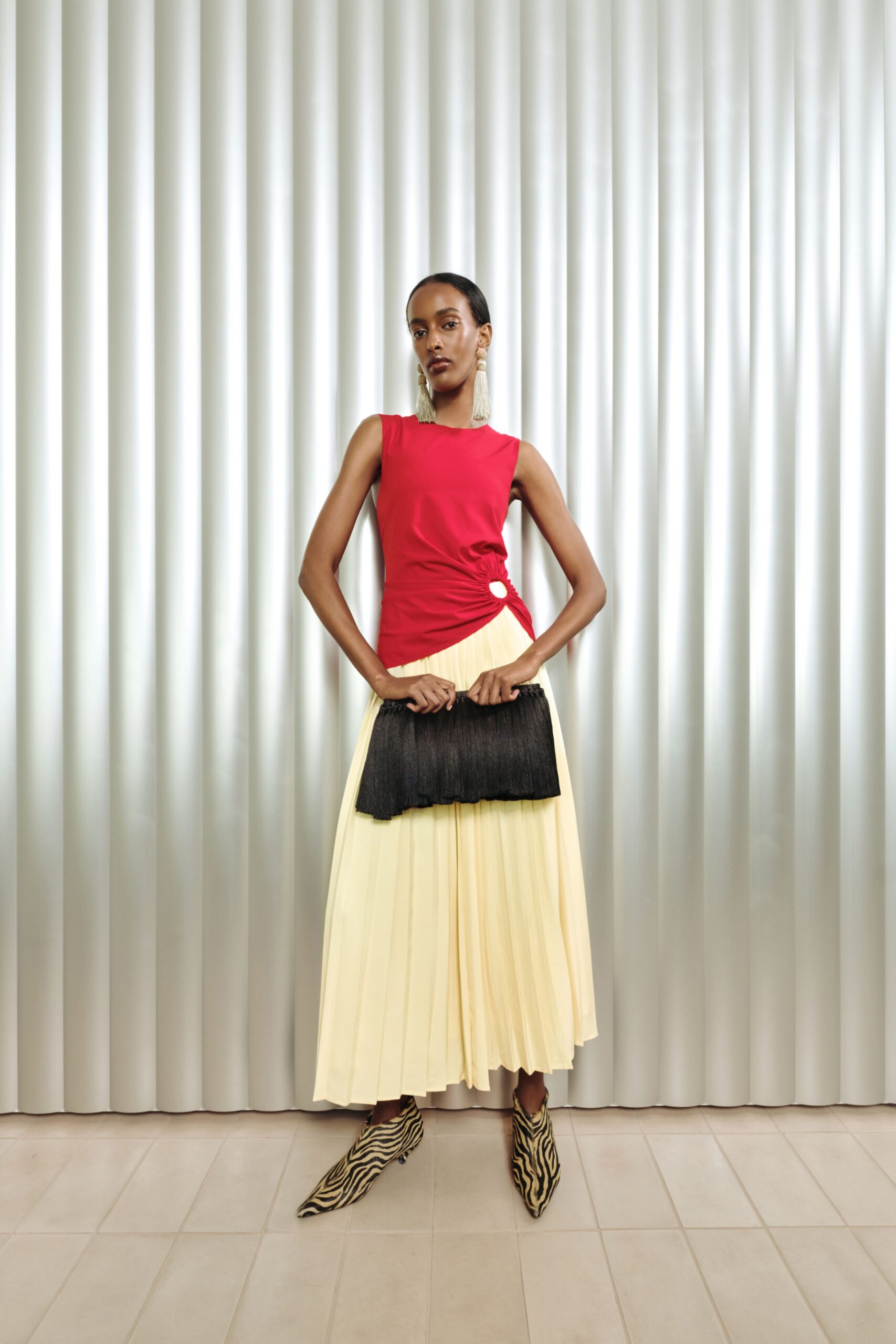 Scarlet sleeveless top ruched into a white ring at the hip, butter-cream skirt in knife pleats. Black crinkled clutch, long tassel earrings, zebra calf-hair pointed boots. Waiting in a hotel lounge before the pianist starts.