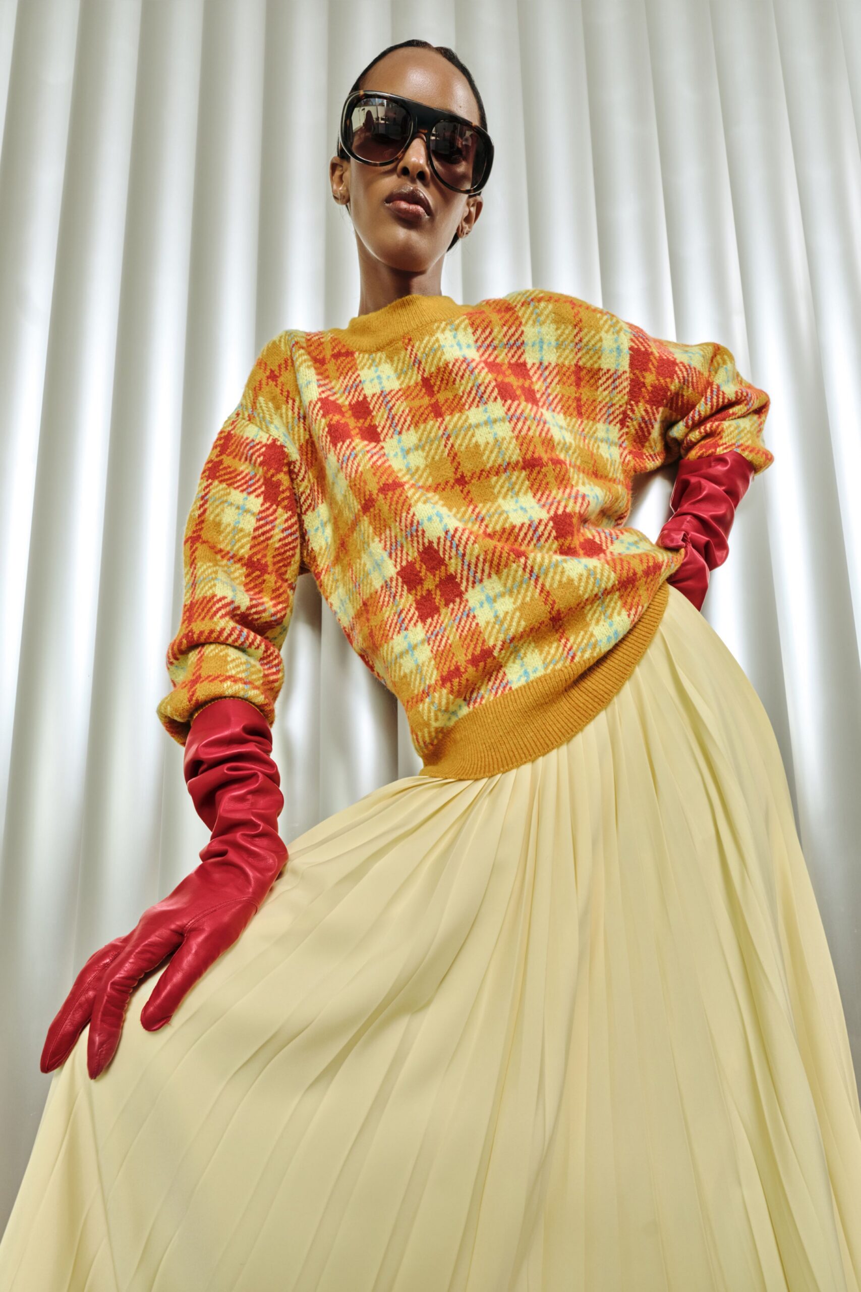 Oversized mustard plaid knit sweater with a ribbed hem sitting low on the hip, opera gloves in cherry leather ruched at the forearm, and a butter-cream pleated skirt. Hotel ballroom curtains, half an hour before speeches.