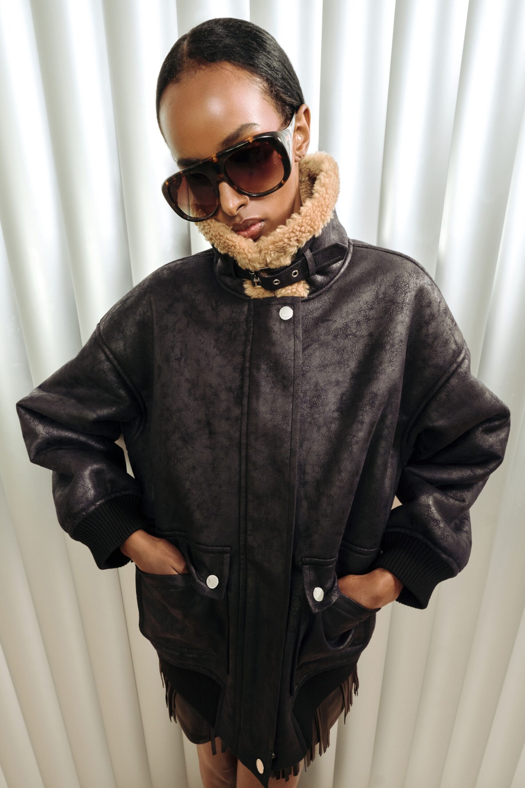 Dark shearling-lined jacket in crackled black leather, the throat latch buckled tight under the chin. Fringed leather skirt peeking below, oversized tortoiseshell aviators. Hotel corridor ice machine humming at 2 a.m.