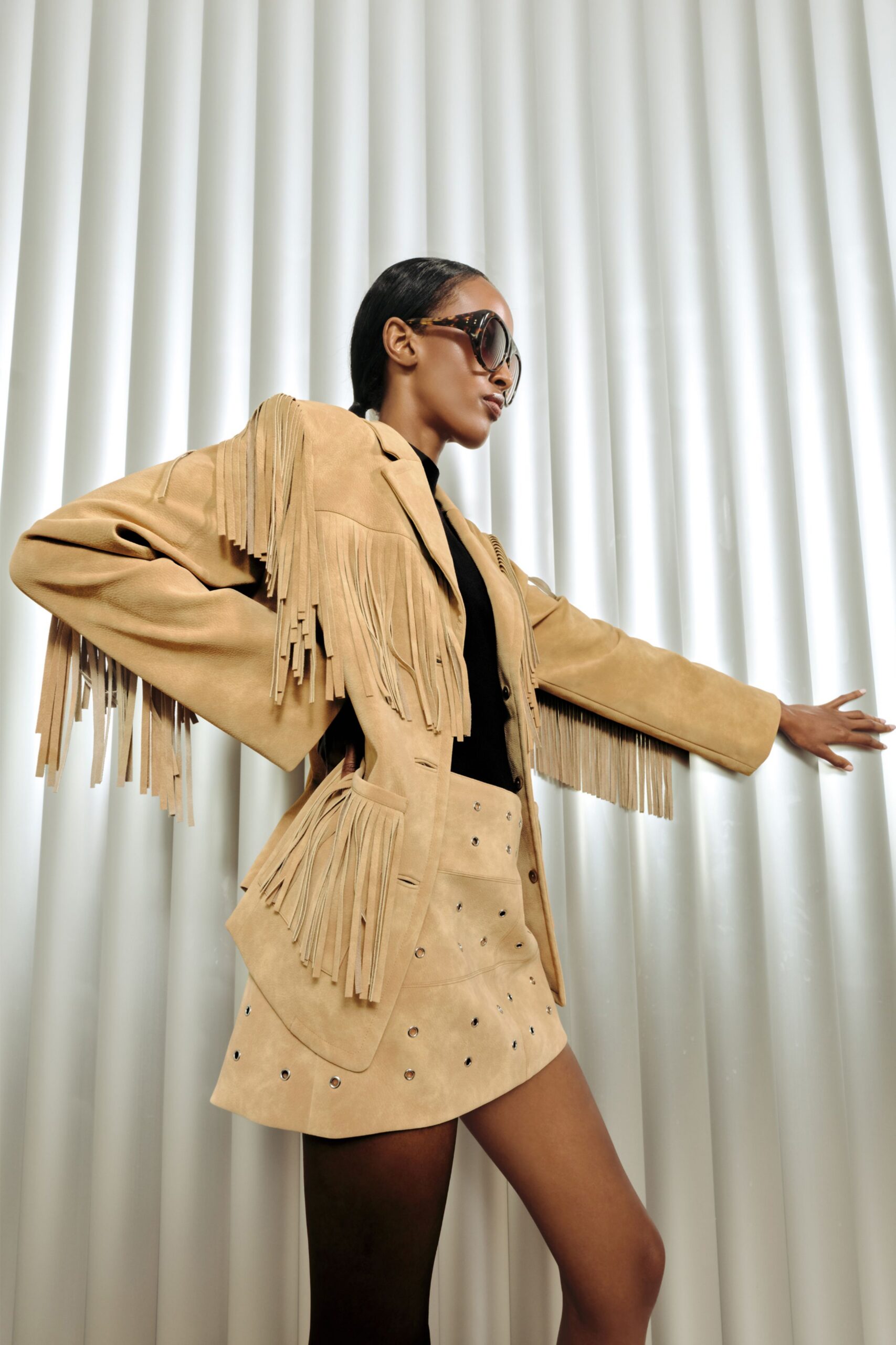 Sand suede blazer with long fringe falling from the yoke and sleeves, over a black high-neck knit. Matching suede mini skirt punched with metal eyelets. Oversized tortoiseshell sunglasses. Waiting outside a desert motel at noon.