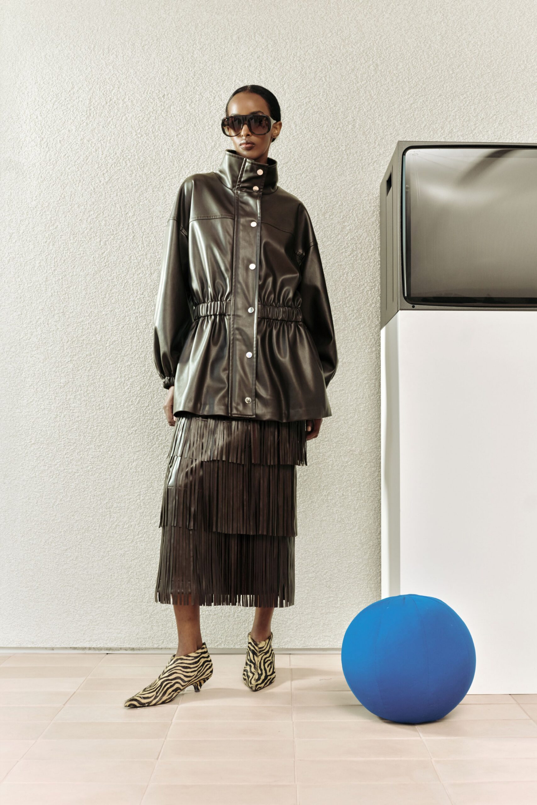 Dark brown leather coat with a stand collar and silver snaps, cinched by a shirred waist. Matching midi skirt cut in tiered fringe. Zebra-print calf hair ankle boots, smoked shield sunglasses. Waiting in a tiled lobby beside a dead TV.