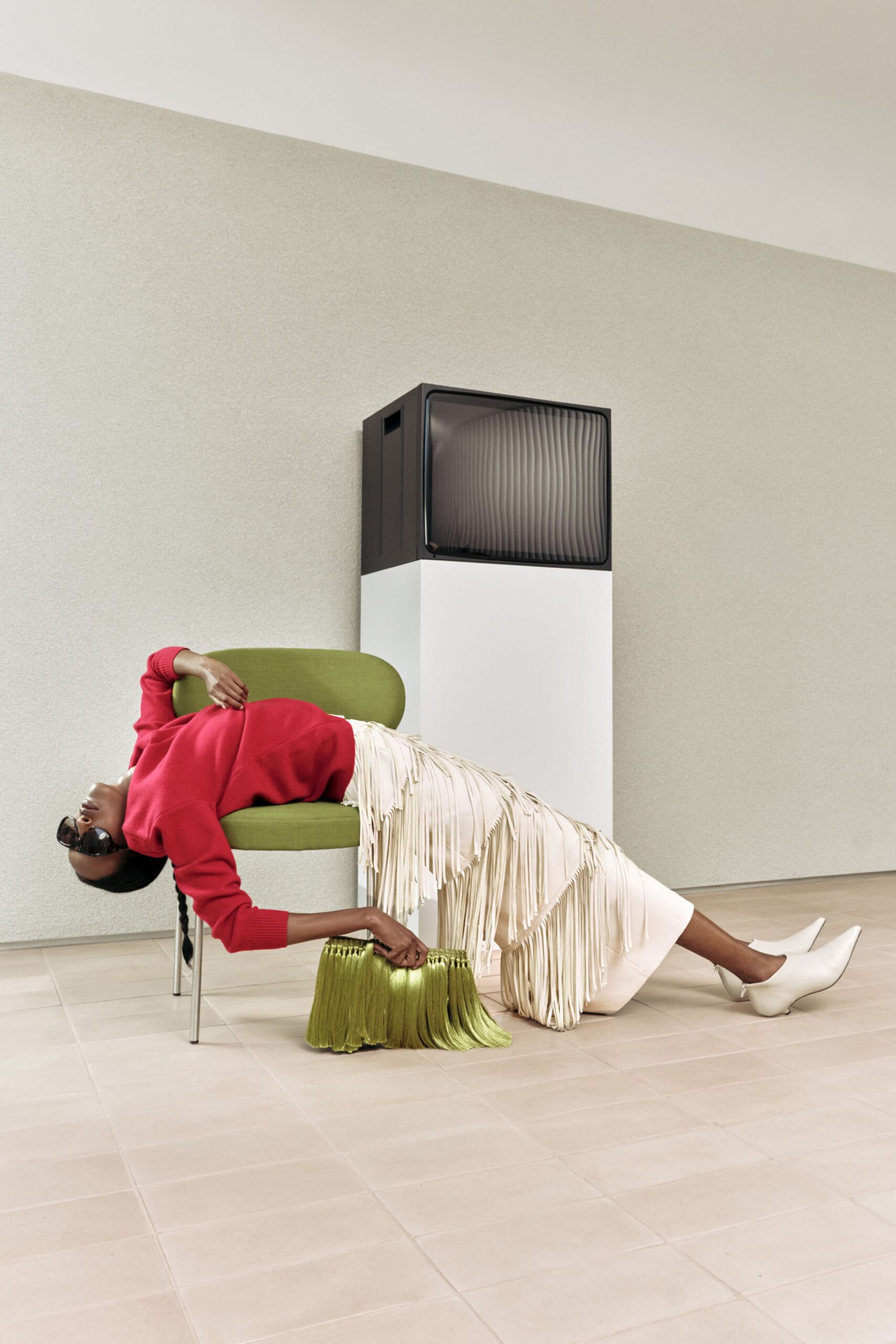 Scarlet rib-knit sweater slouched off one shoulder, ivory leather skirt banded with long swaying fringe tiers. White pointed ankle boots with a curved heel, black sunglasses, pleated chartreuse bag. Waiting out a museum lobby lull.
