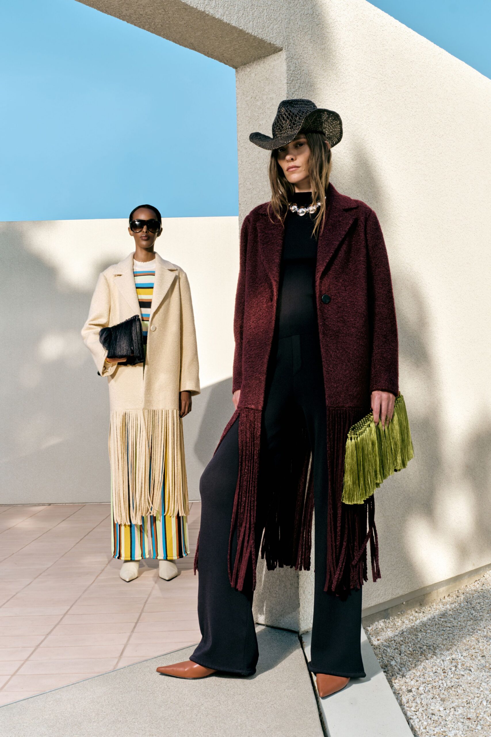 Open-weave black cowboy hat, aubergine bouclé coat with long fringe swinging from the hem, black knit top, fluid black trousers breaking over tan pointed boots. Olive tassel clutch in hand. Noon on a stucco terrace gone quiet.