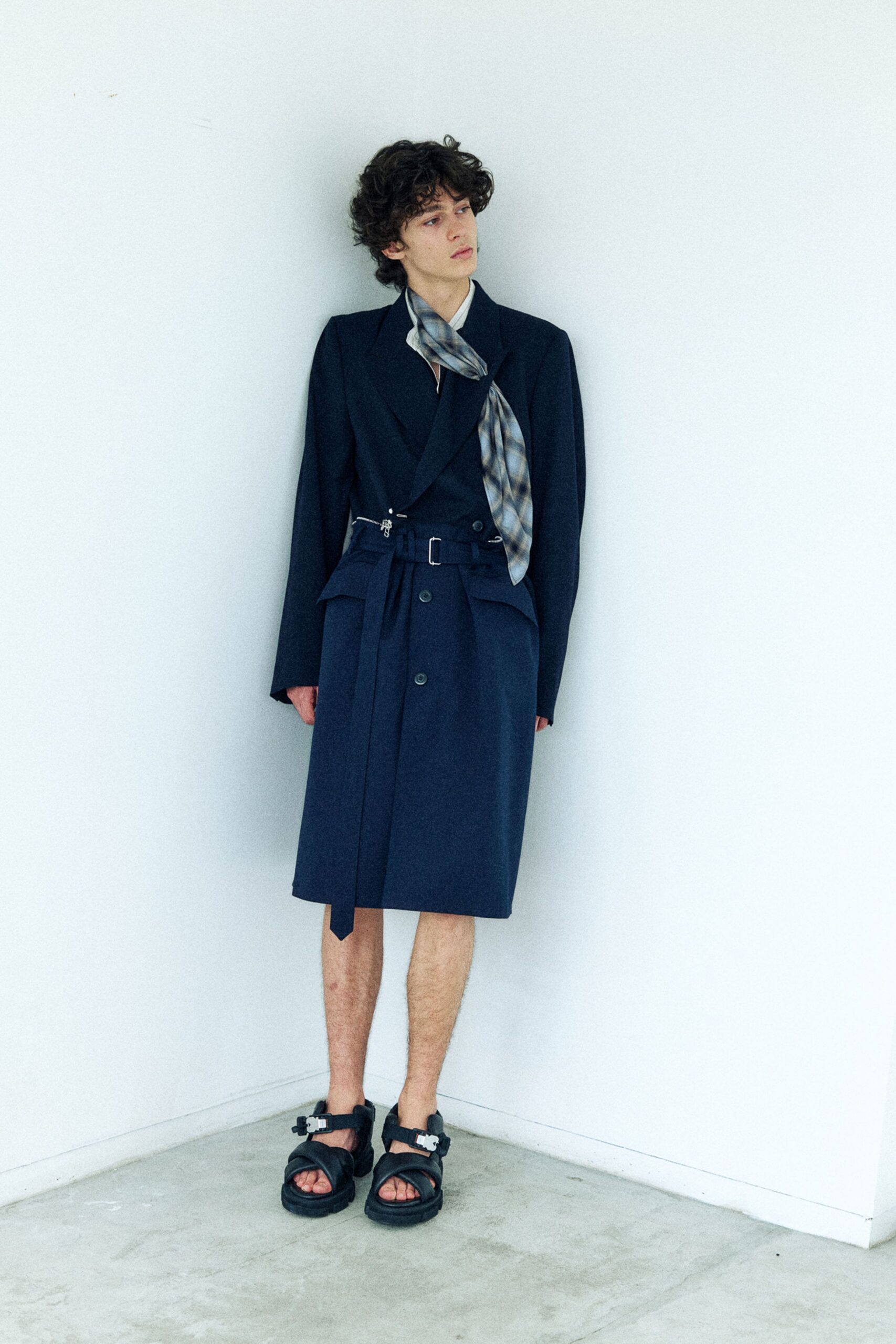 Dark wool blazer cut long through the sleeve, a smoky plaid scarf knotted loose at the throat. Matching wrap skirt with a hanging belt tab and clipped waist chain, black trekking sandals. Waiting out a museum lobby draft.
