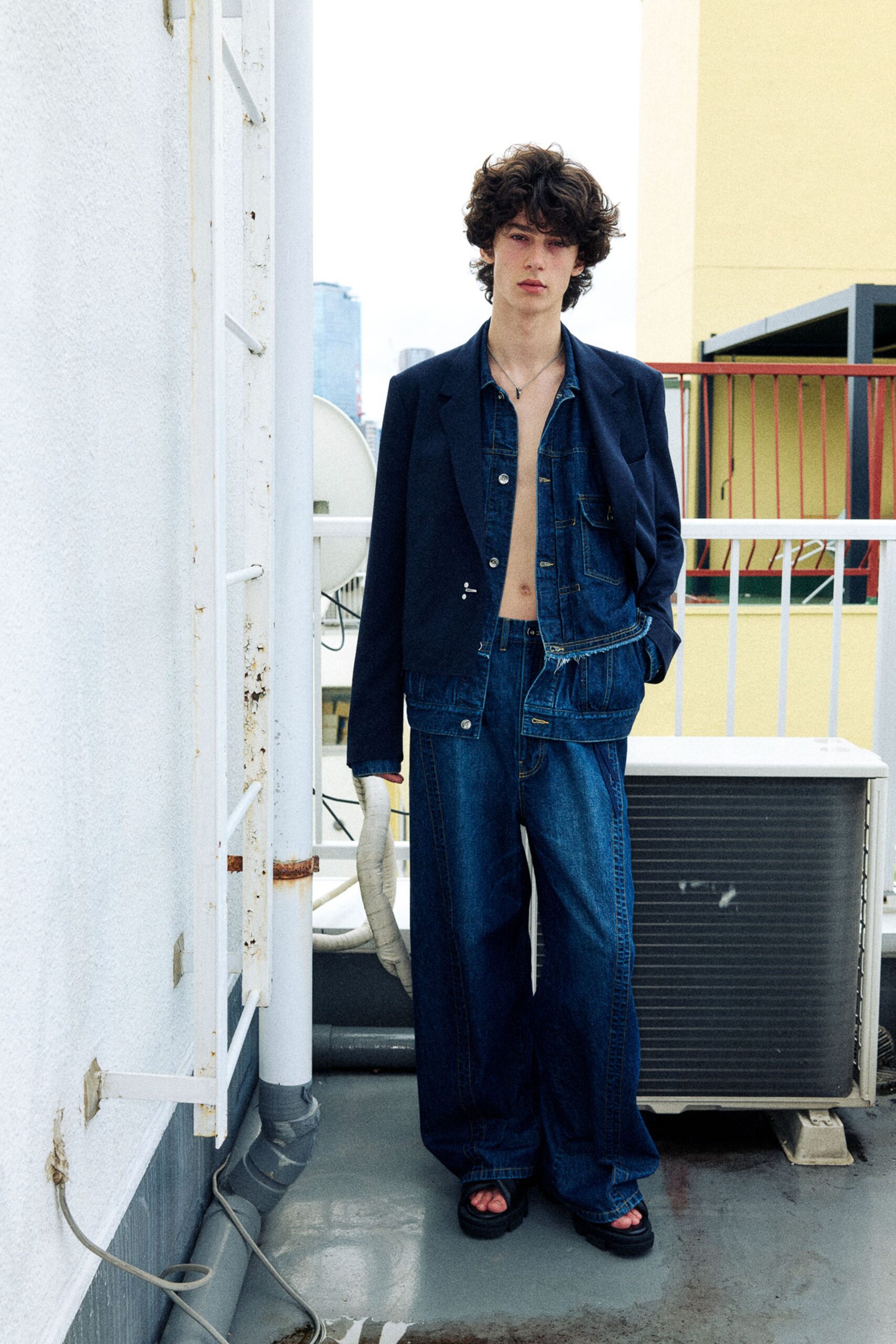Navy wool blazer with a low silver clasp, dark indigo denim shirt left open with a frayed hem, matching wide-leg jeans with deep curved seaming, black slide sandals. On a damp rooftop beside the humming air unit.
