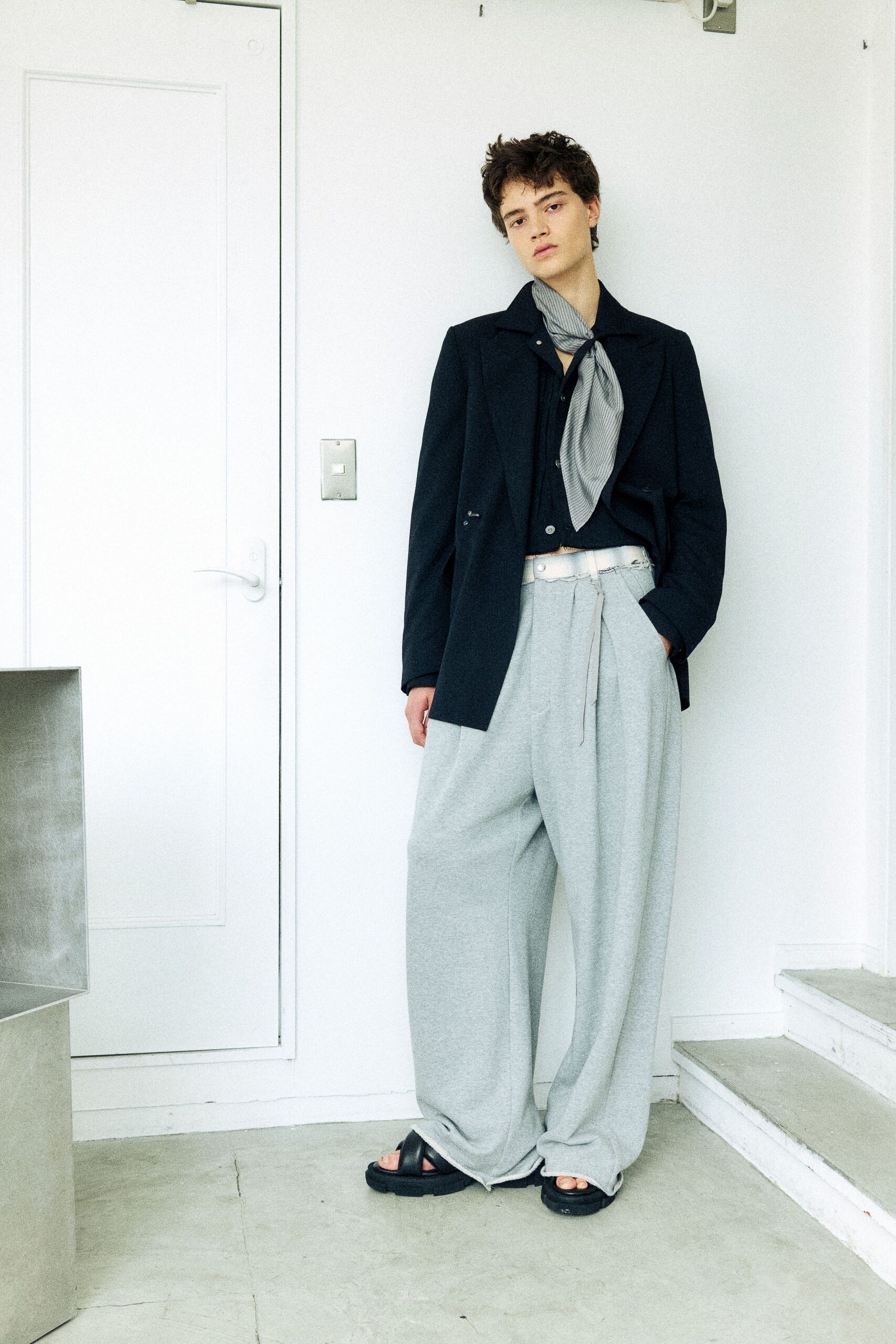 Black blazer with notch lapels over a black shirt fastened with pearly buttons, a grey pinstriped scarf knotted loose at the throat. Pale heather wide-leg trousers with a raw waistband, black slide sandals. Waiting by a side door after the lift jams.