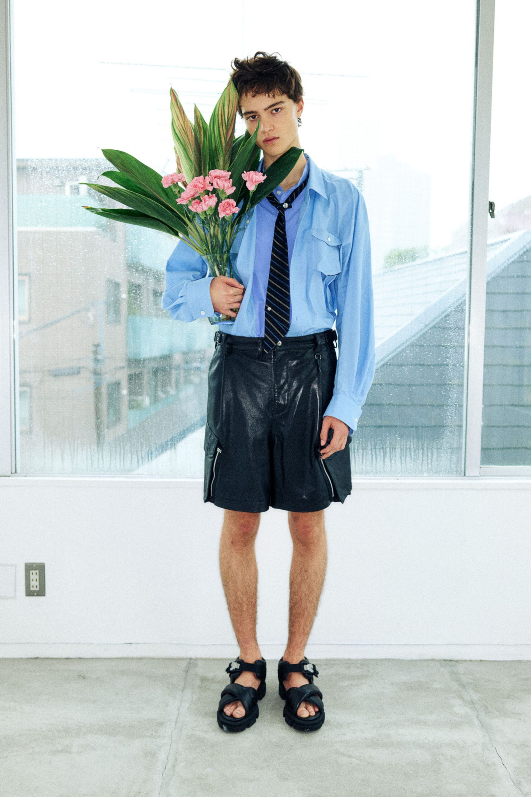 Pale blue button-down shirt with a flap pocket snapped in white, navy striped tie pinned with a pearl bead, black leather shorts with zip cargo pockets, thick strap sandals. Rain on the window, flowers in hand after a missed apology.