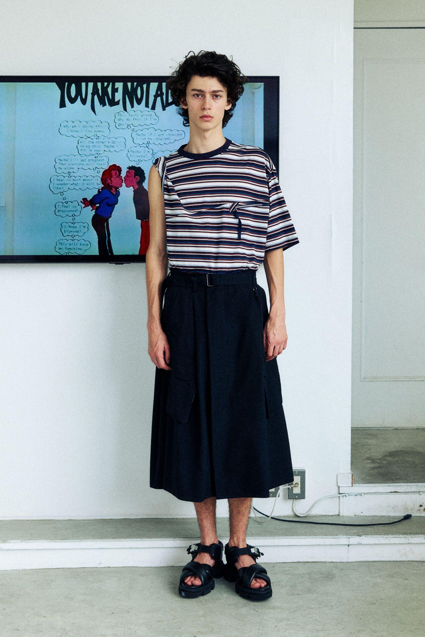 Striped jersey tee cut with one bare armhole and a flap pocket stitched across the chest. Ink-black culottes in dense twill, belted at the waist with hanging cargo tabs. Thick-soled sandals. Waiting out a humid afternoon in an apartment gallery.
