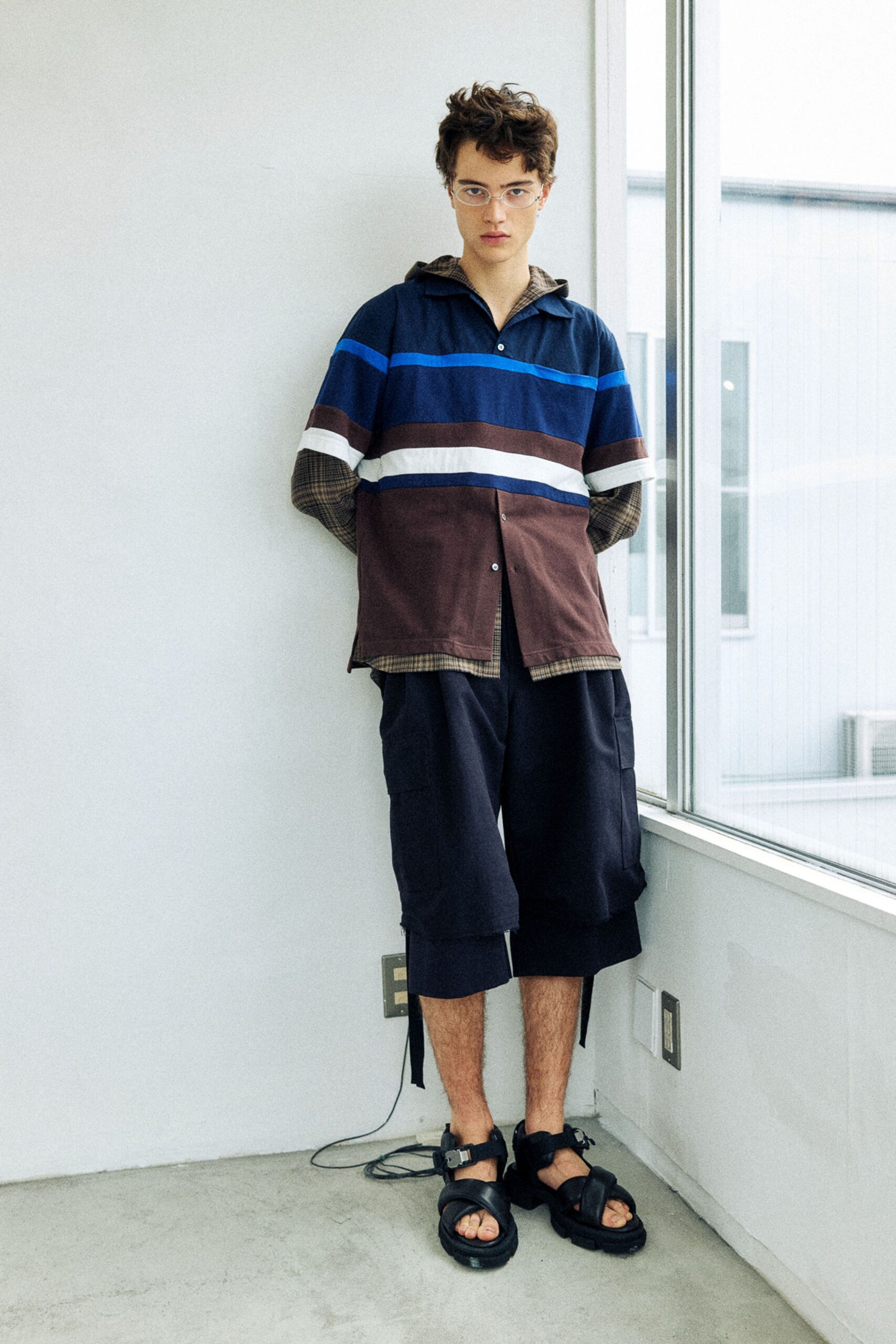 Navy rugby shirt with electric-blue and white chest bands, layered over a brown micro-check hooded shirt peeking past the hem. Cropped navy cargo shorts with long hanging tabs, black buckle sandals. By the office window after everyone else has left.