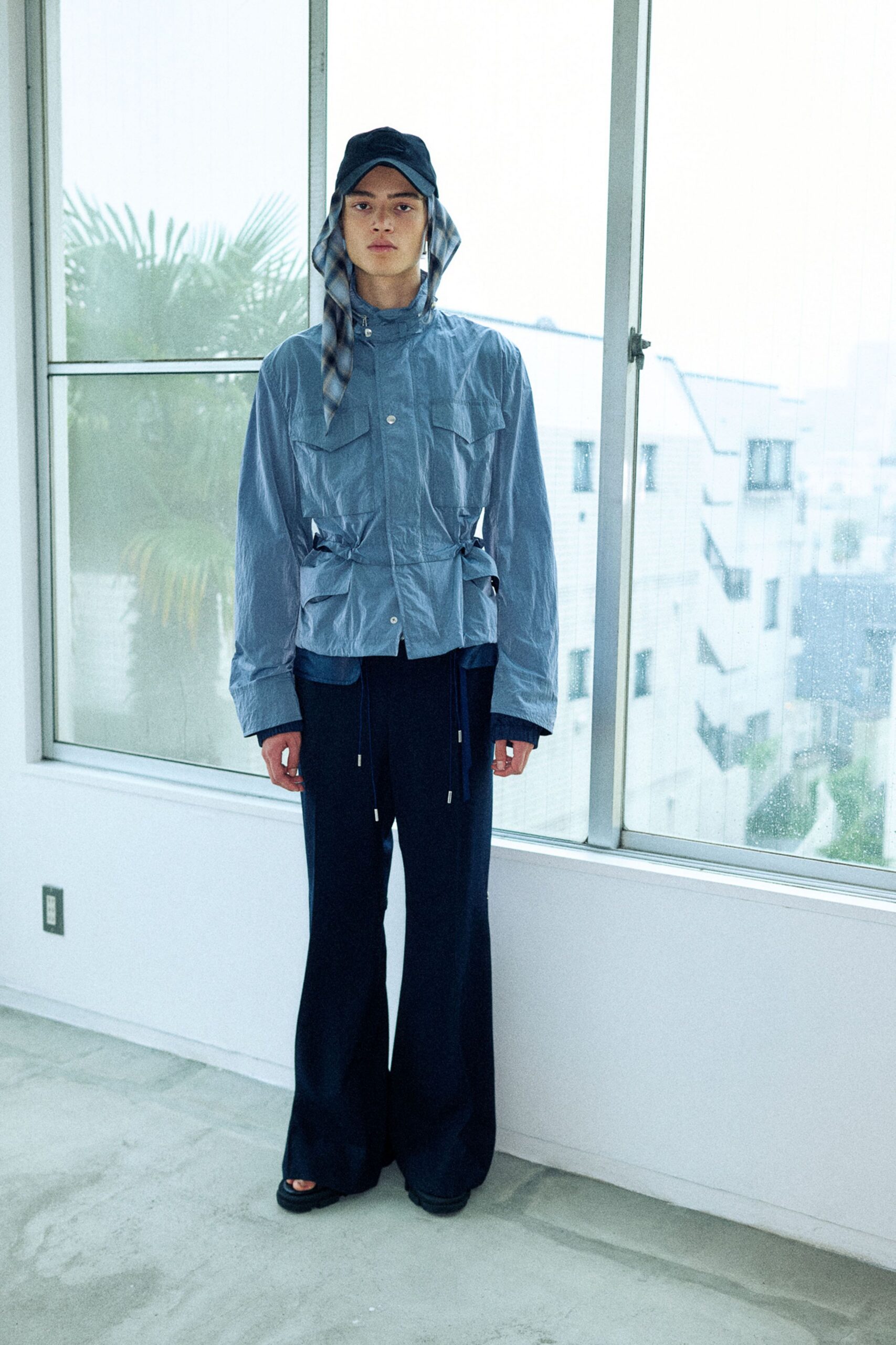 Black baseball cap with a checked scarf tucked beneath it. Slate crinkled nylon field jacket with flap pockets and cinched side tabs, dark flared trousers with long drawstrings, black slide sandals. Rain beading on a high-rise window.