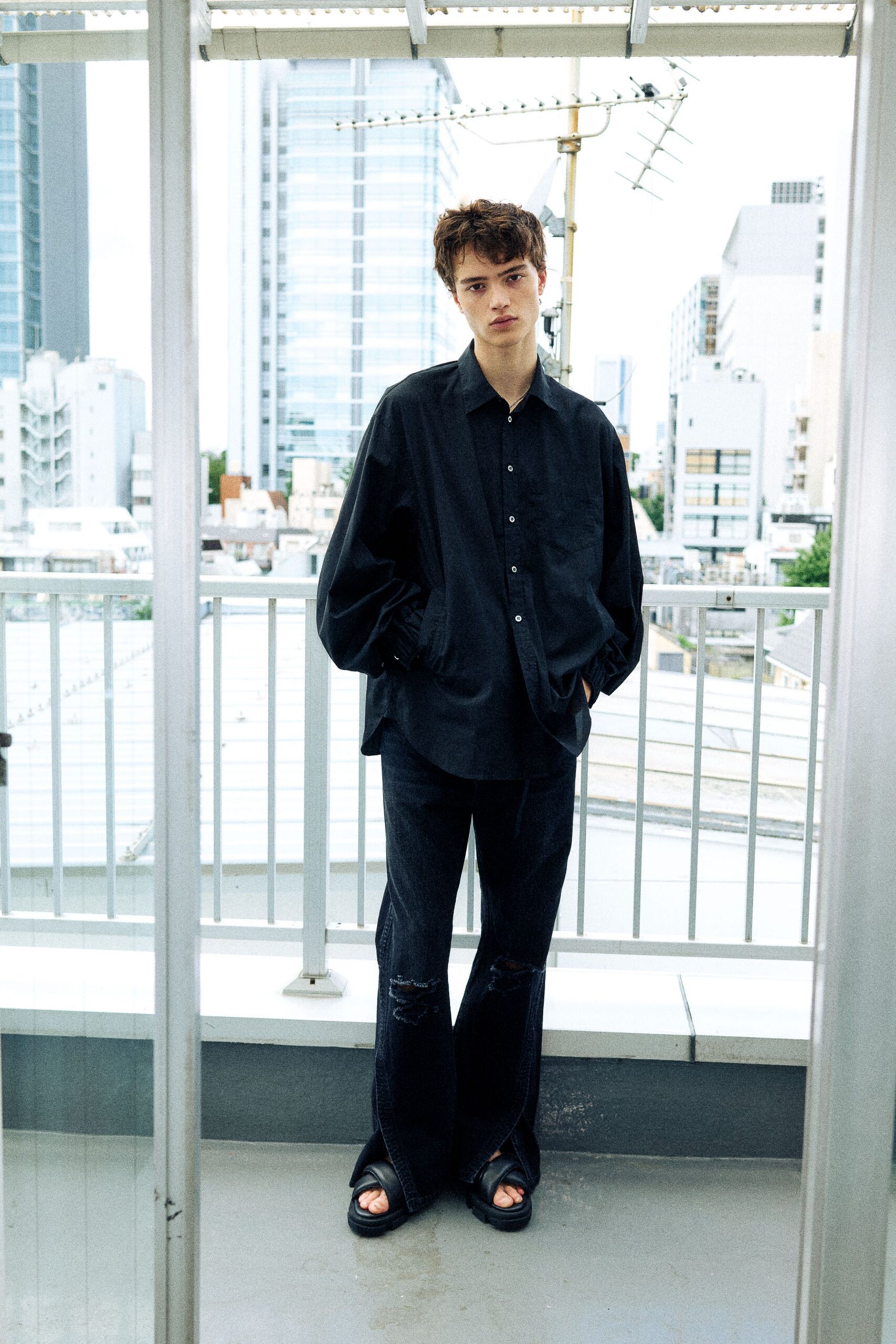 Oversized black shirt in crisp poplin, the placket skewed slightly off center. Faded black jeans with torn knees and a loose break over chunky leather slides. On a rooftop walkway, waiting out the heat after rain.