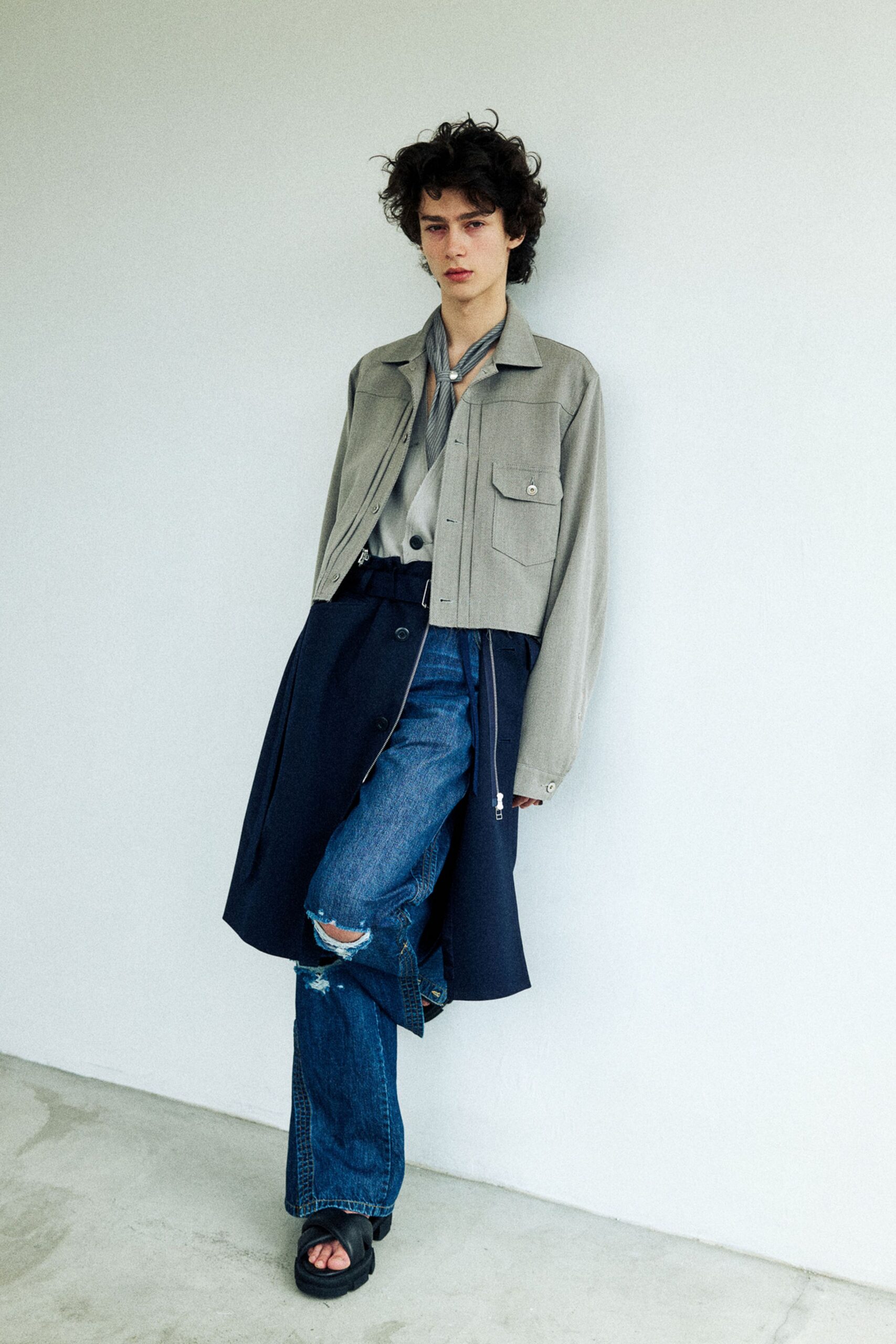 Stone cropped shirt jacket with flap pocket and a zip hem, over a draped dove camisole and narrow striped neck scarf pinned with a pearl. Inky wrap skirt panel over ripped blue jeans, black slides. Waiting out drizzle under a station awning.