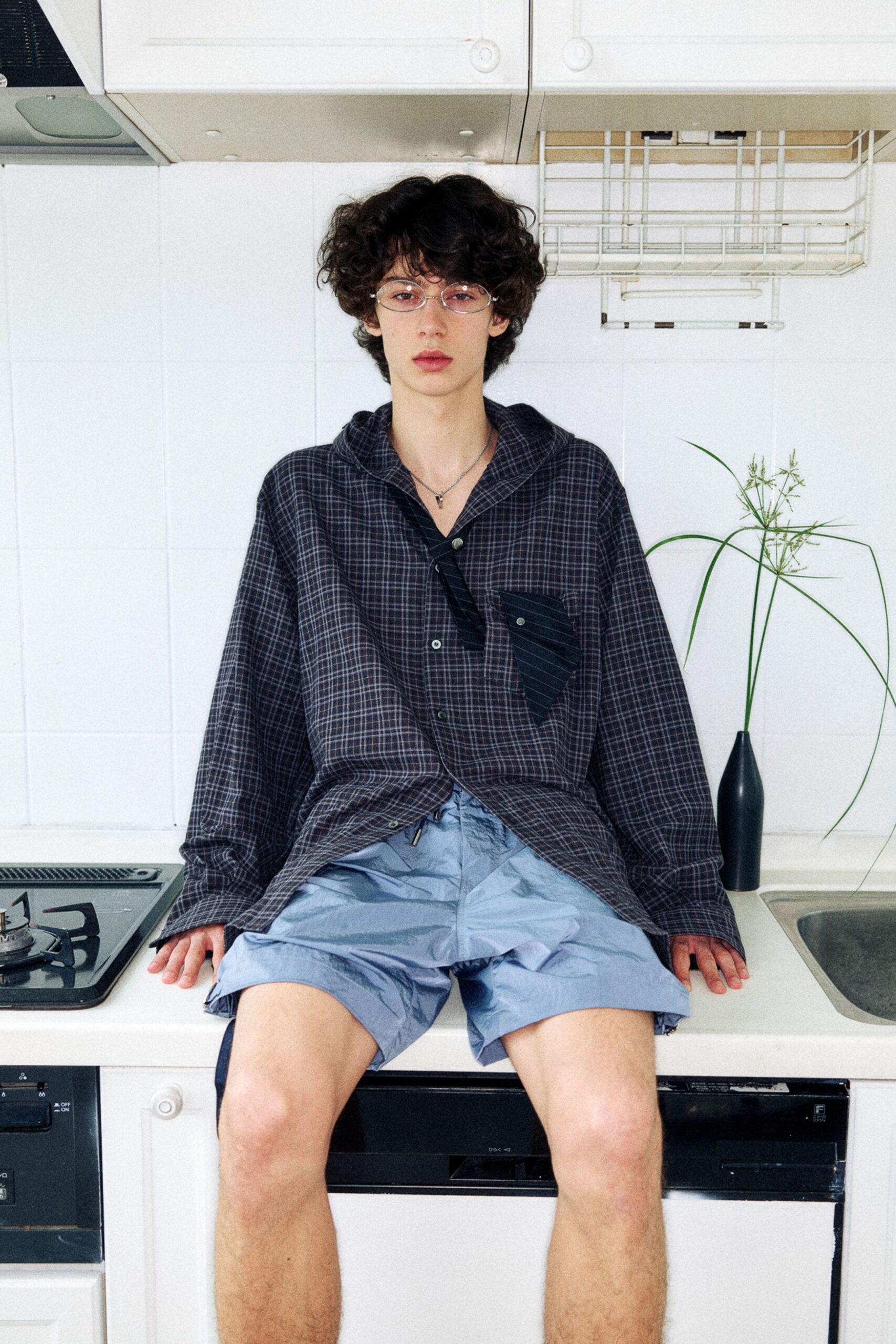 Charcoal windowpane hooded shirt with a mismatched pinstripe placket and chest pocket, mother-of-pearl buttons undone low. Pale blue nylon shorts with a drawcord knot peeking out. Perched on the kitchen counter after rinsing peaches.