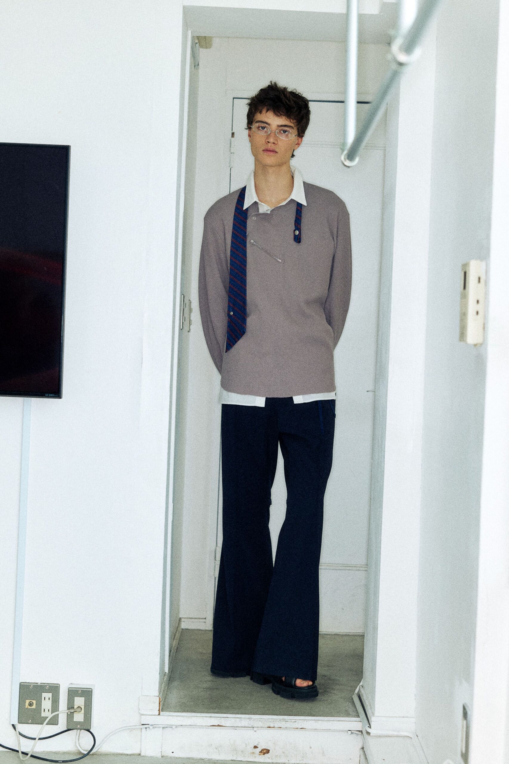 Taupe fine-rib knit pullover pinned at the chest, over a white poplin shirt with the hem hanging below. Navy flared trousers with a sharp crease, black slide sandals. In a narrow apartment hall, waiting for the buzzer.
