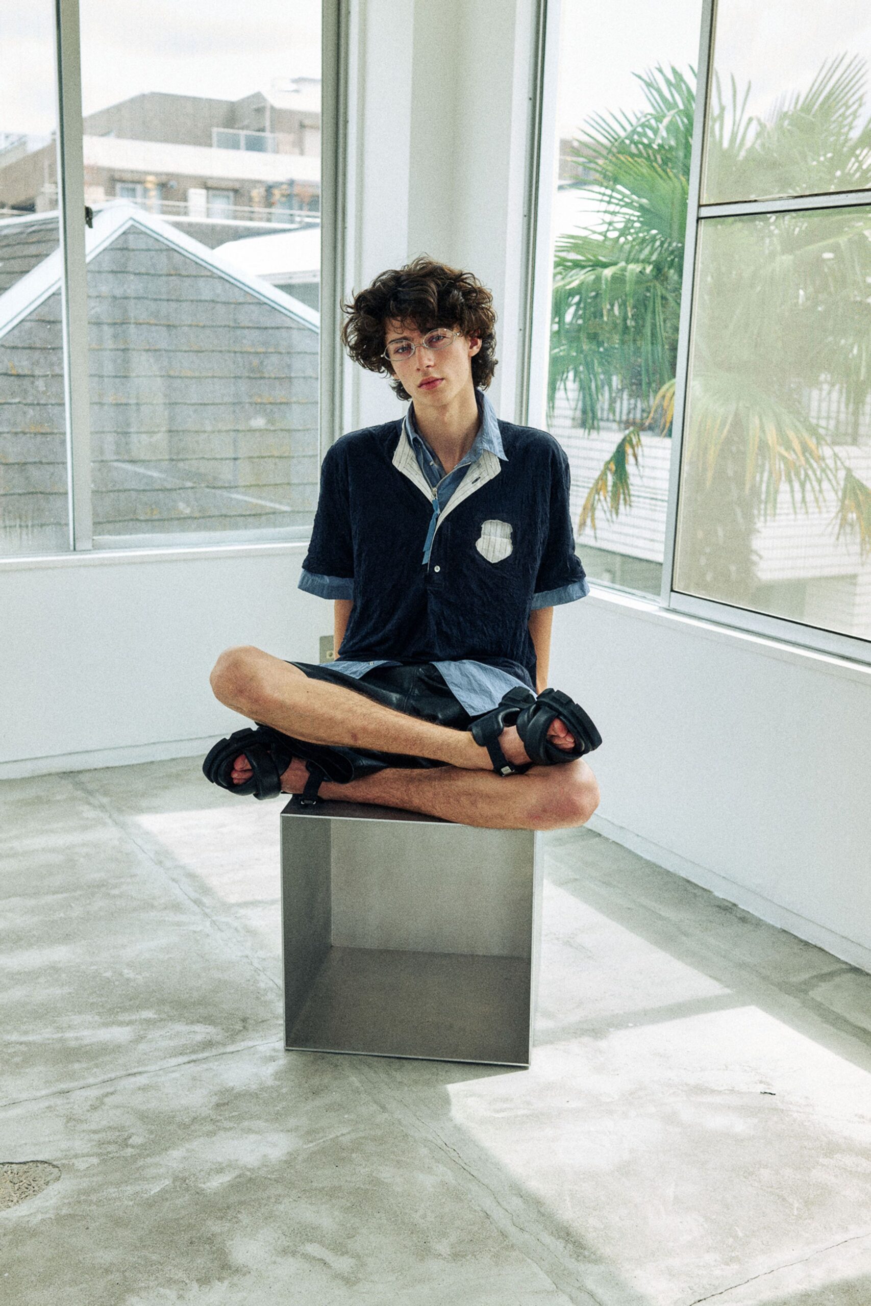 Navy crinkled short-sleeve shirt with a white pinstriped patch pocket, layered over a pale chambray shirt left hanging past the hem. Black leather shorts, padded strap sandals. Sunlight on a concrete floor after the windows have been opened.