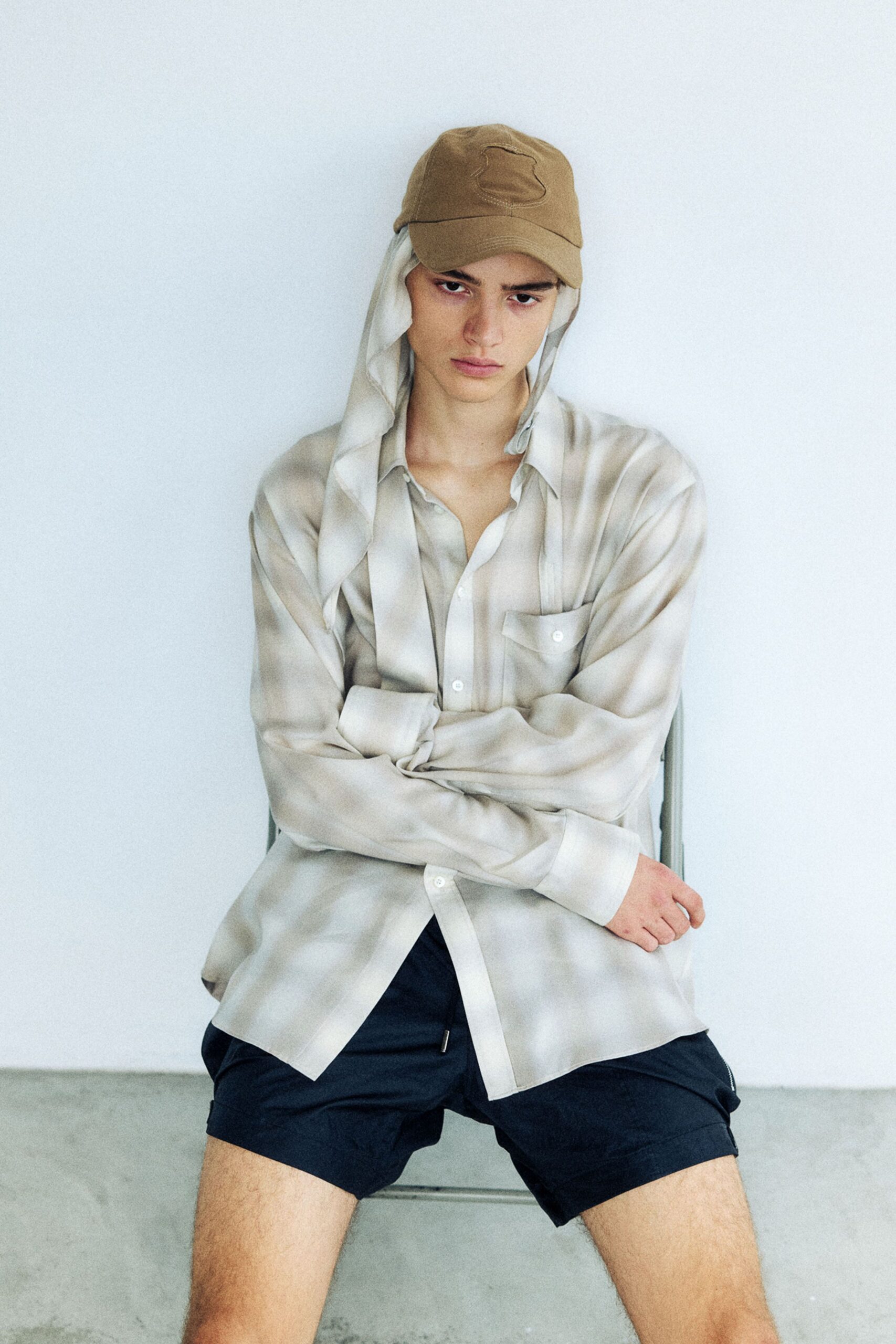 Tan twill cap with a long ivory scarf tucked under it, pale stone shirt in fluid satin with a flap chest pocket, black nylon shorts cut high on the thigh. Waiting out the heat in a bare rehearsal room.