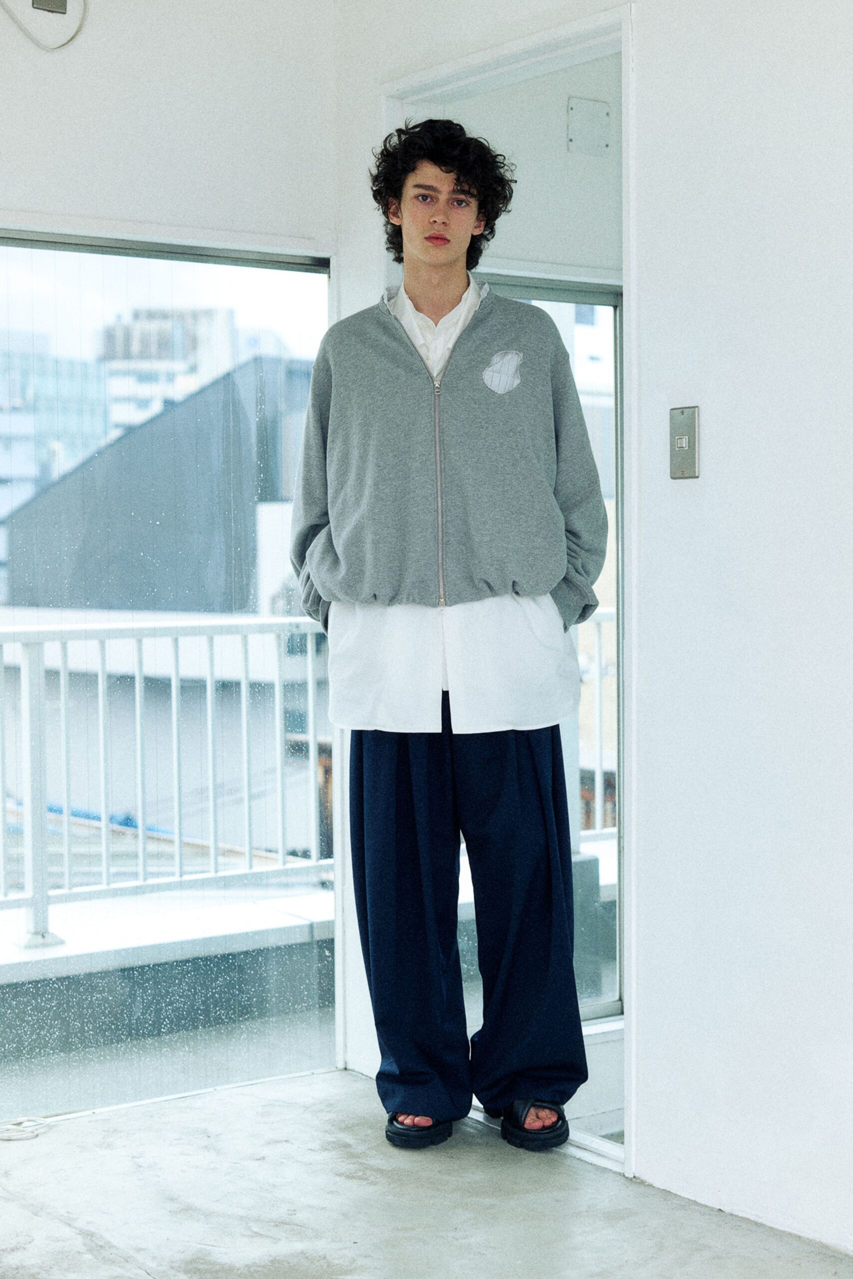Heather-grey jersey zip jacket with a quilted chest patch, over a white band-collar shirt cut long past the hem. Ink-black wide trousers pooling at the sandals. Waiting by the balcony door on a washed-out morning.