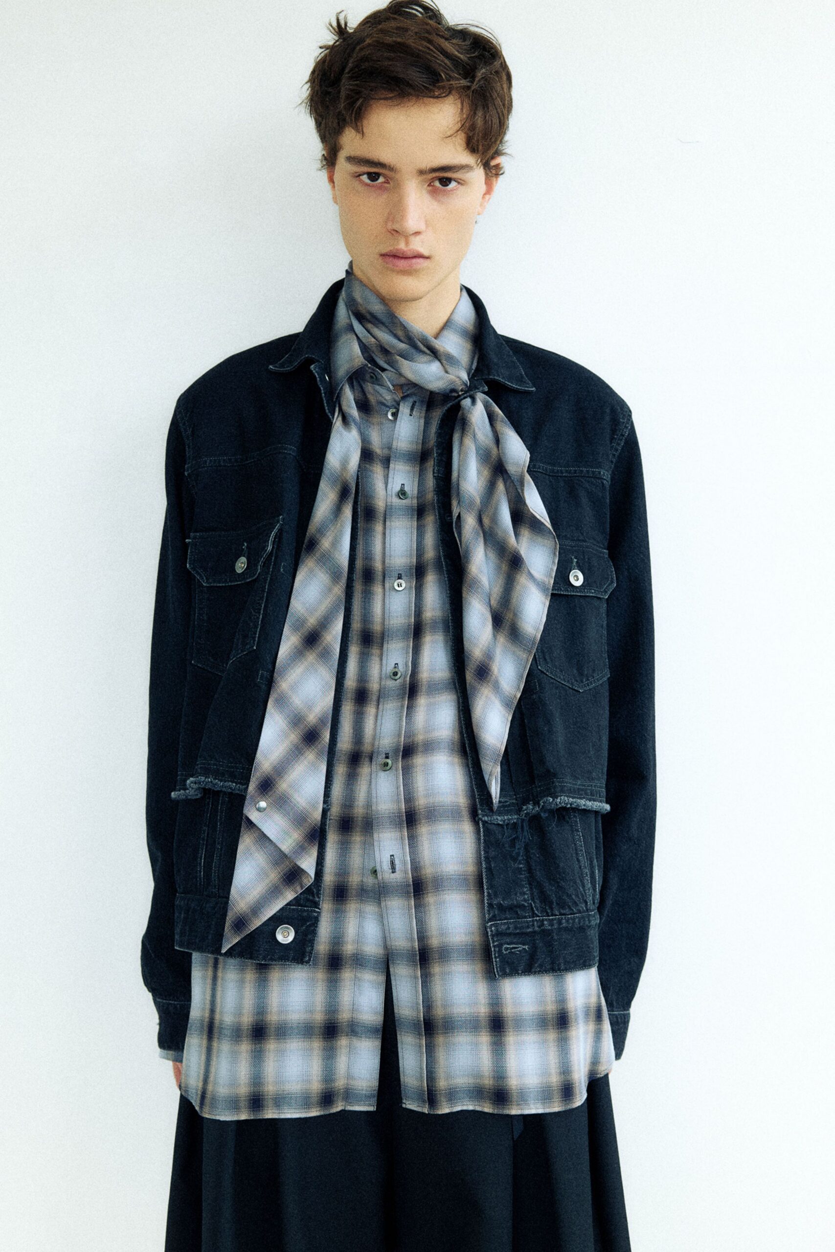 Cropped black denim jacket with a raw, uneven hem over a misty plaid shirt dress with pearly grey buttons, the matching plaid scarf knotted loose at the throat. Like waiting in a station cafe after the last train's gone.