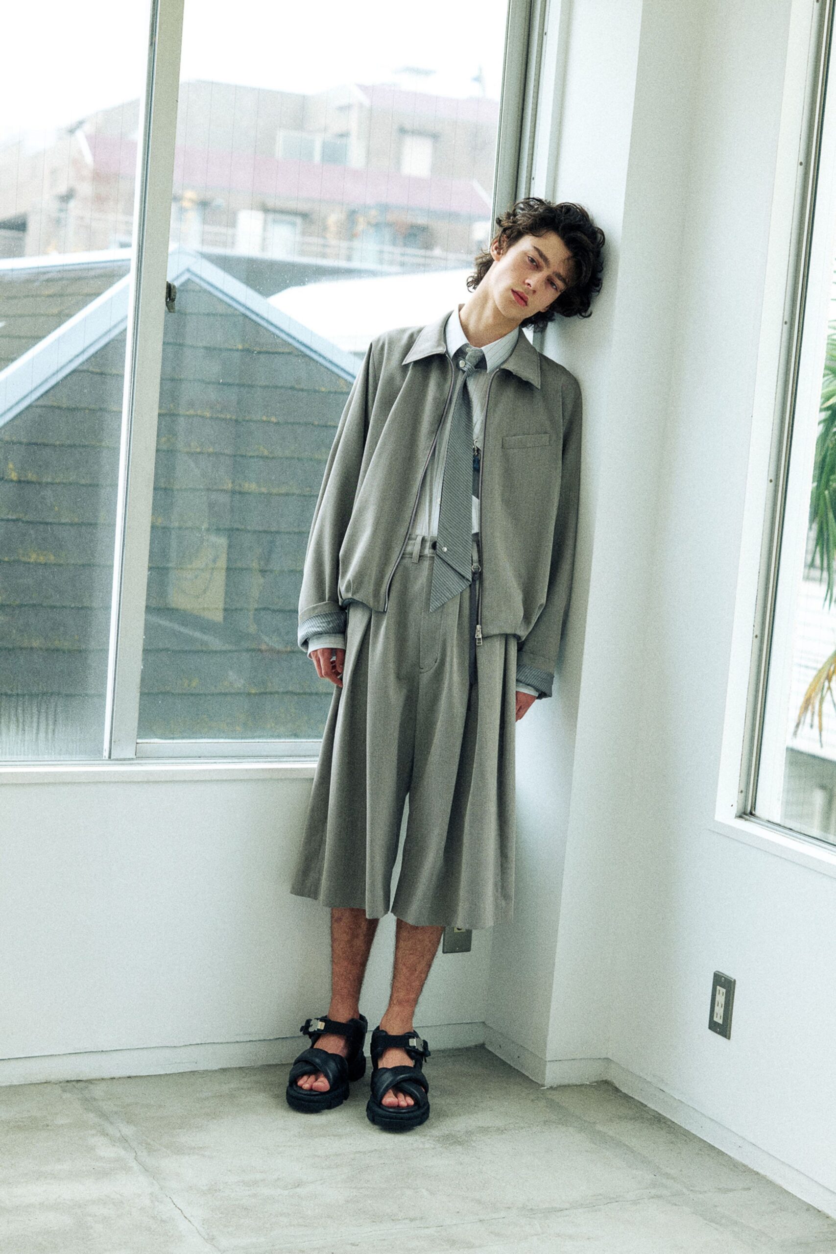 Pale sage zip-front jacket with a dropped shoulder, striped poplin shirt and narrow tie, gauzy culottes falling from a double-tab waistband, black sandals with padded straps. Leaning by sun-warmed glass after a late checkout.