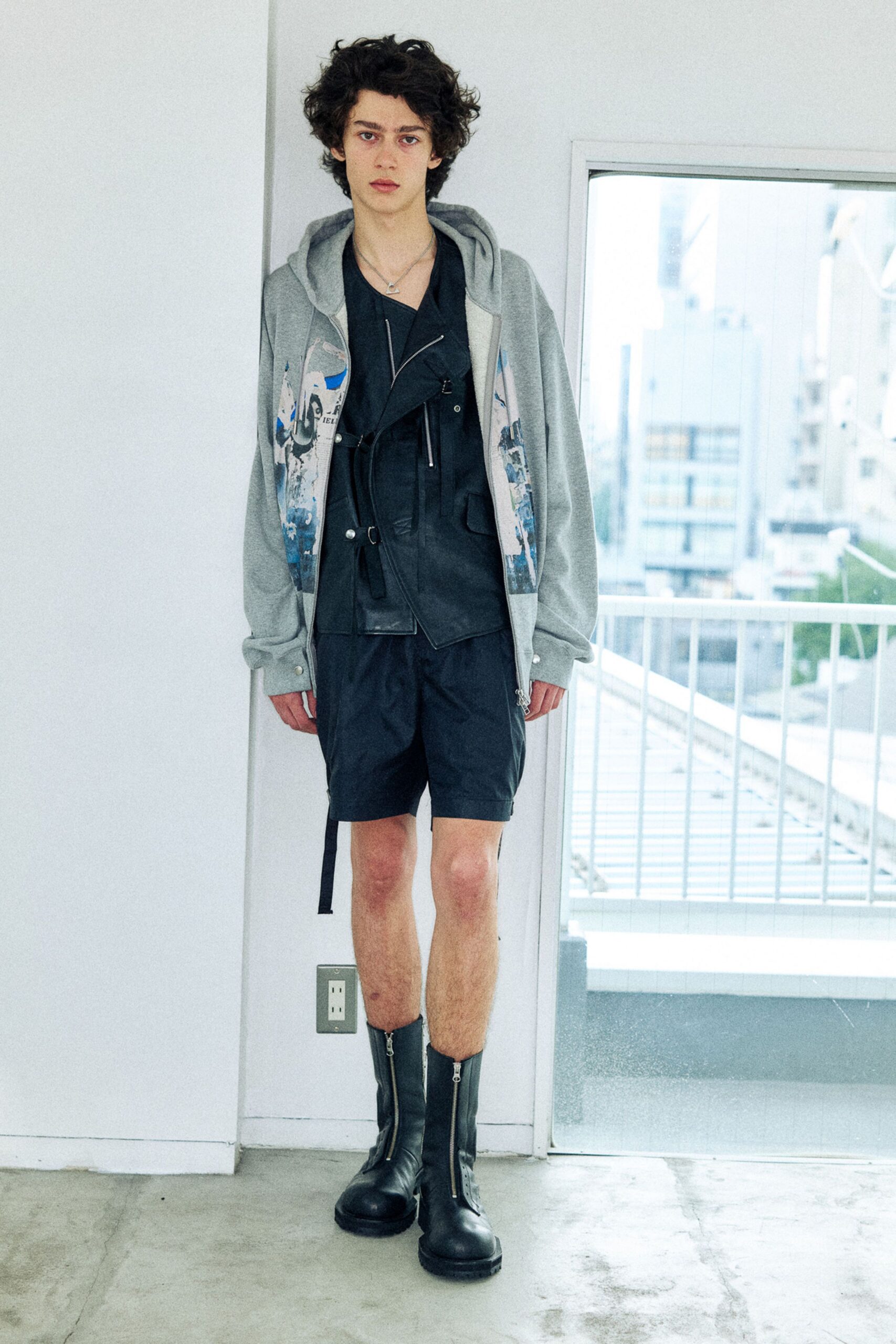 Grey jersey zip hoodie with washed photo panels, over a black wrap vest traced with exposed zips and a dangling strap. Black nylon shorts, front-zip leather ankle boots. Waiting for the service lift to rattle open.