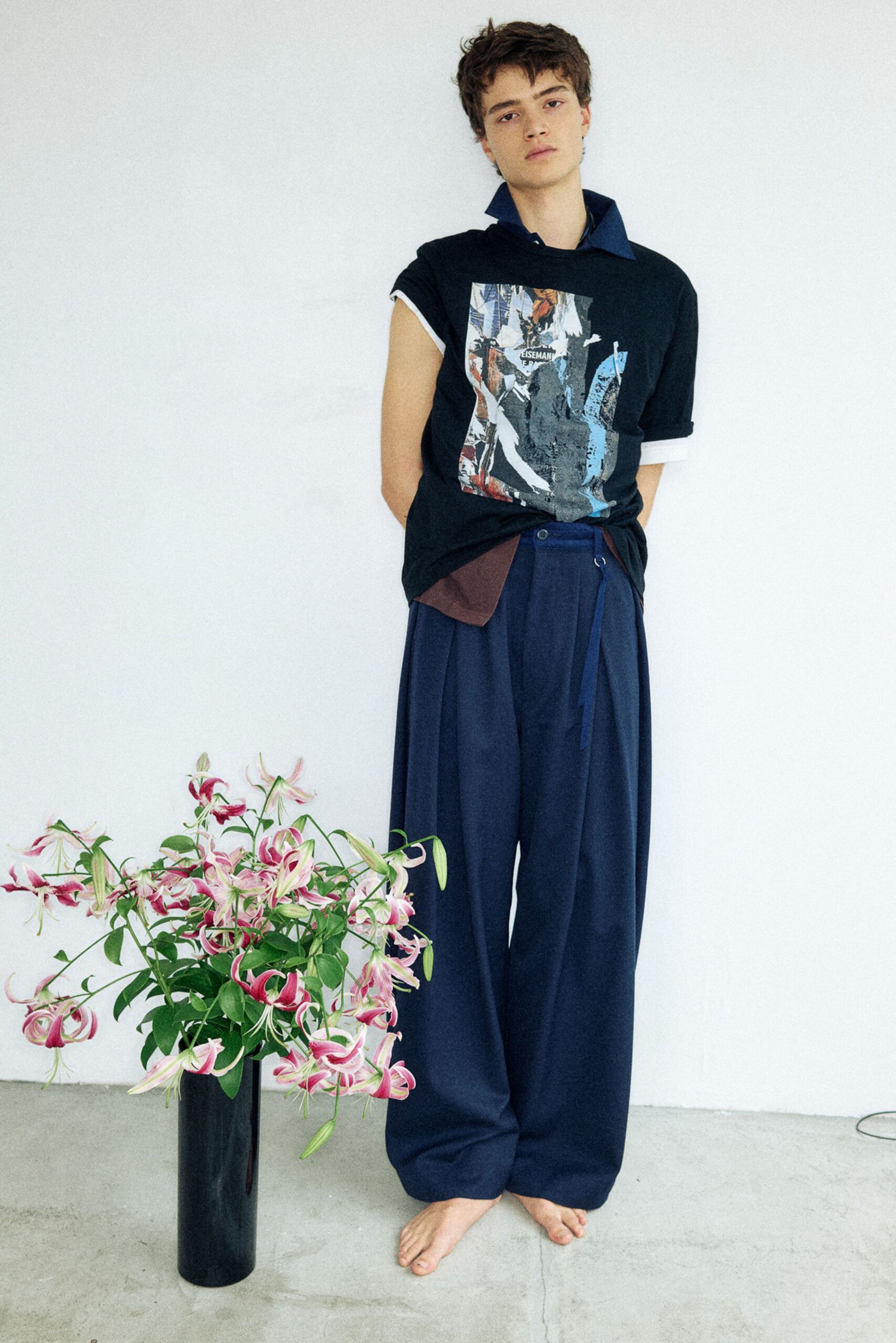 Black T-shirt with a pasted-collage print and turned-up cuffs over a navy shirt with a sharp spread collar. Ink-blue wide-leg trousers fall from deep pleats, one belt tab hanging loose. Bare feet by a vase of wilting lilies.