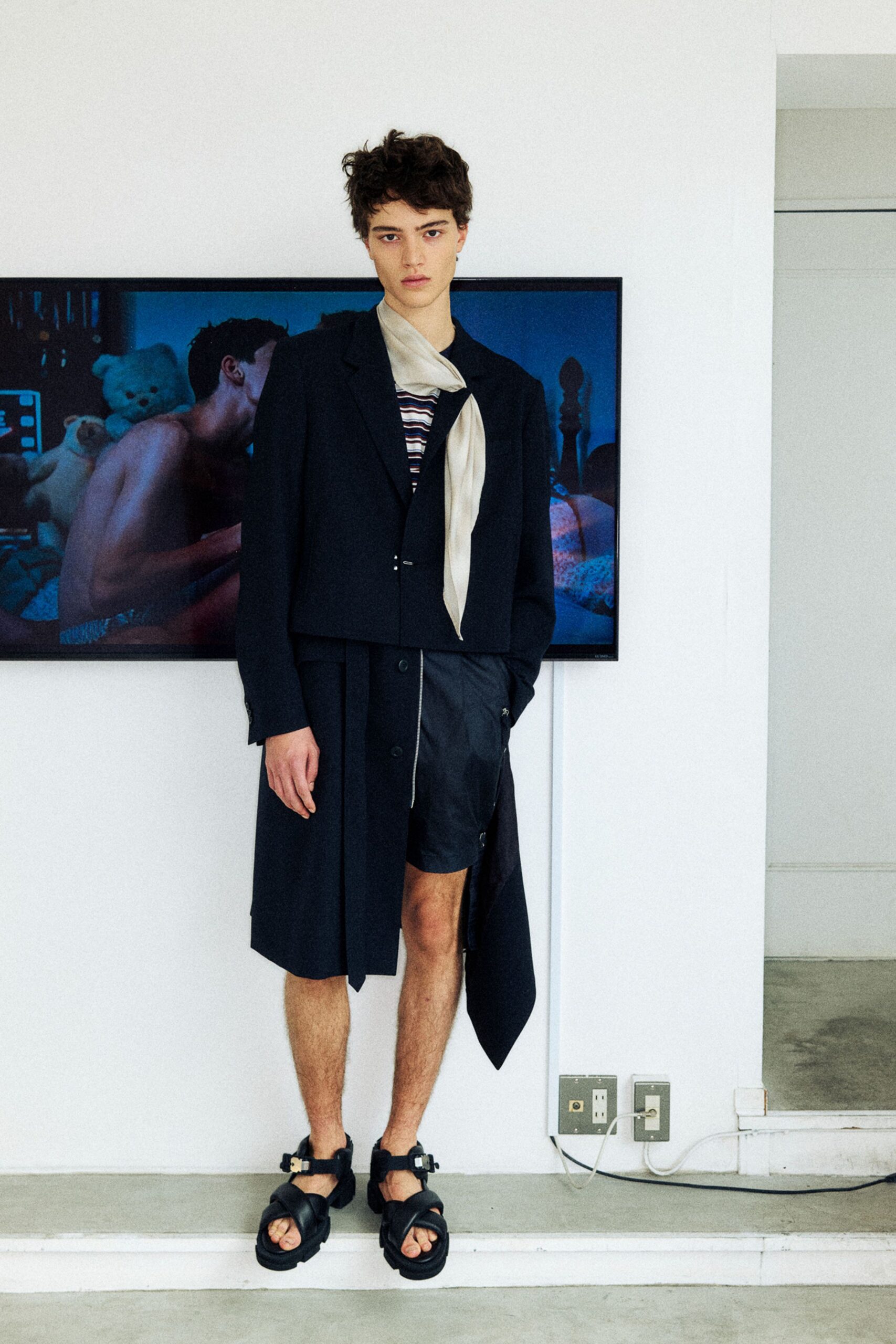 Cropped black blazer with a single hook closure, cream scarf draped loose at the neck, striped knit tee beneath. Black wrap shorts with a dangling tie panel, thick buckle sandals. Waiting in a half-furnished loft while the screen still glows blue.