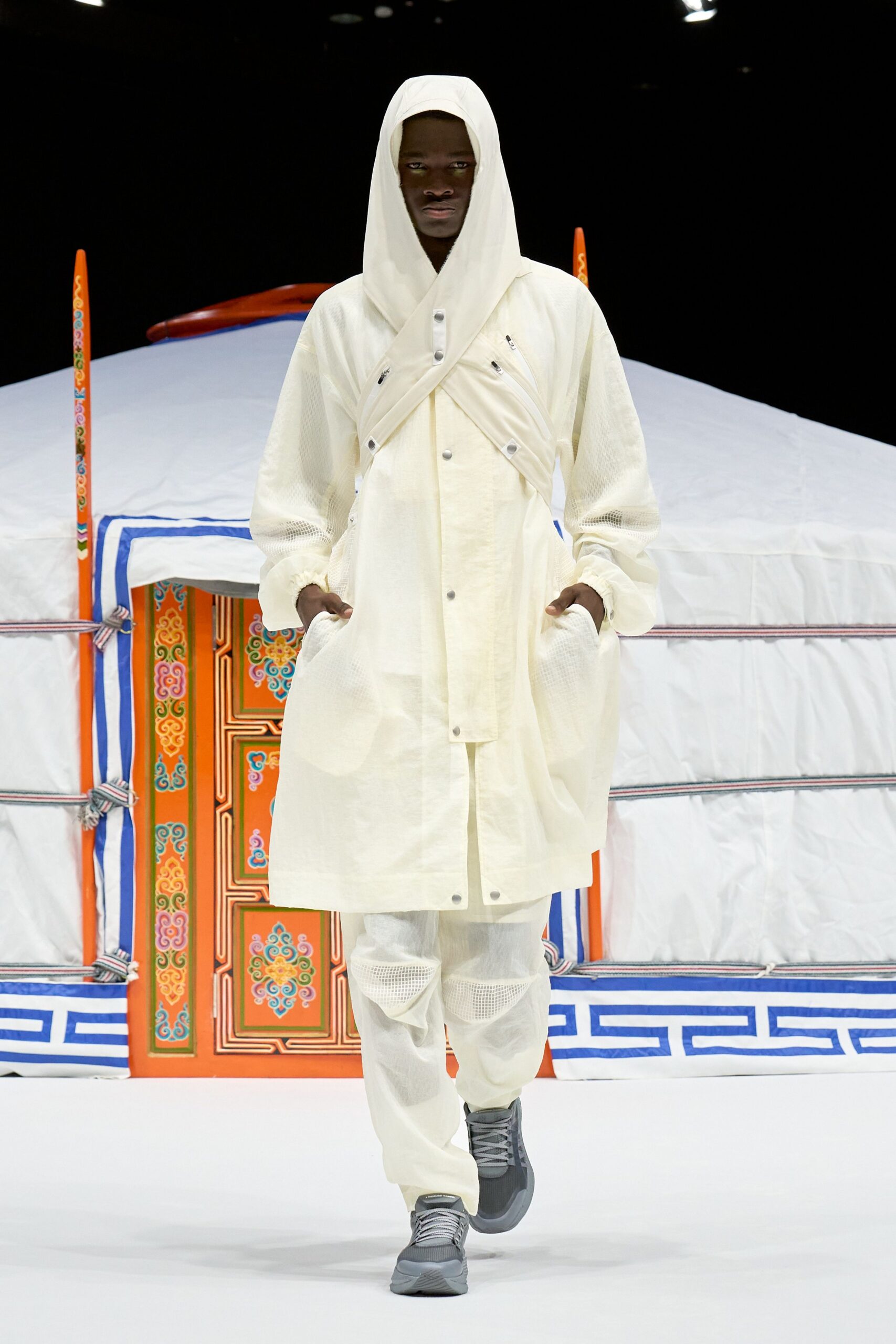 Ivory hooded coat in translucent ripstop, crossed with zippered straps and matte snaps. Matching gauze trousers with mesh knee panels. Gray technical sneakers. Outside a yurt at first light, breath still hanging in the air.
