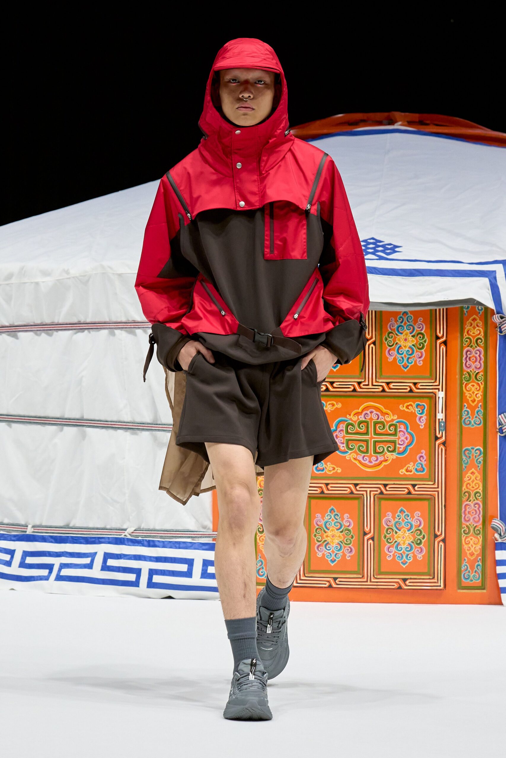 Scarlet and charcoal hooded anorak in crisp shell, cut with diagonal waterproof zips and a snap placket. Black wide shorts in dense jersey, grey trainers. Like stepping out to tighten tent ropes before dawn rain.