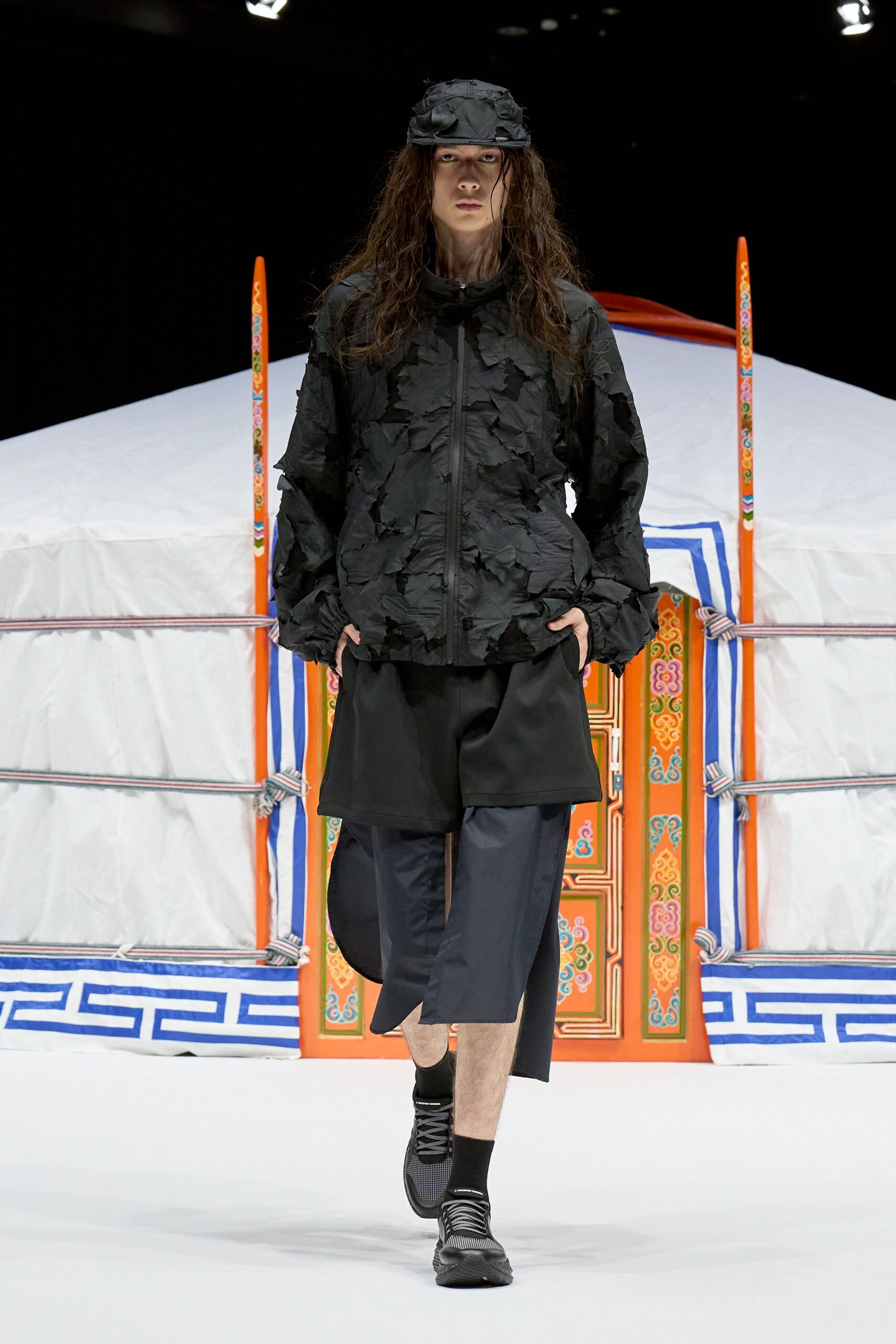 Black cap with frayed appliqué patches, black zip-front jacket in crinkled nylon scattered with raw-edged overlays. Wide black shorts over a split-skirt layer, mesh-toe trainers. Outside a yurt before the rain starts.