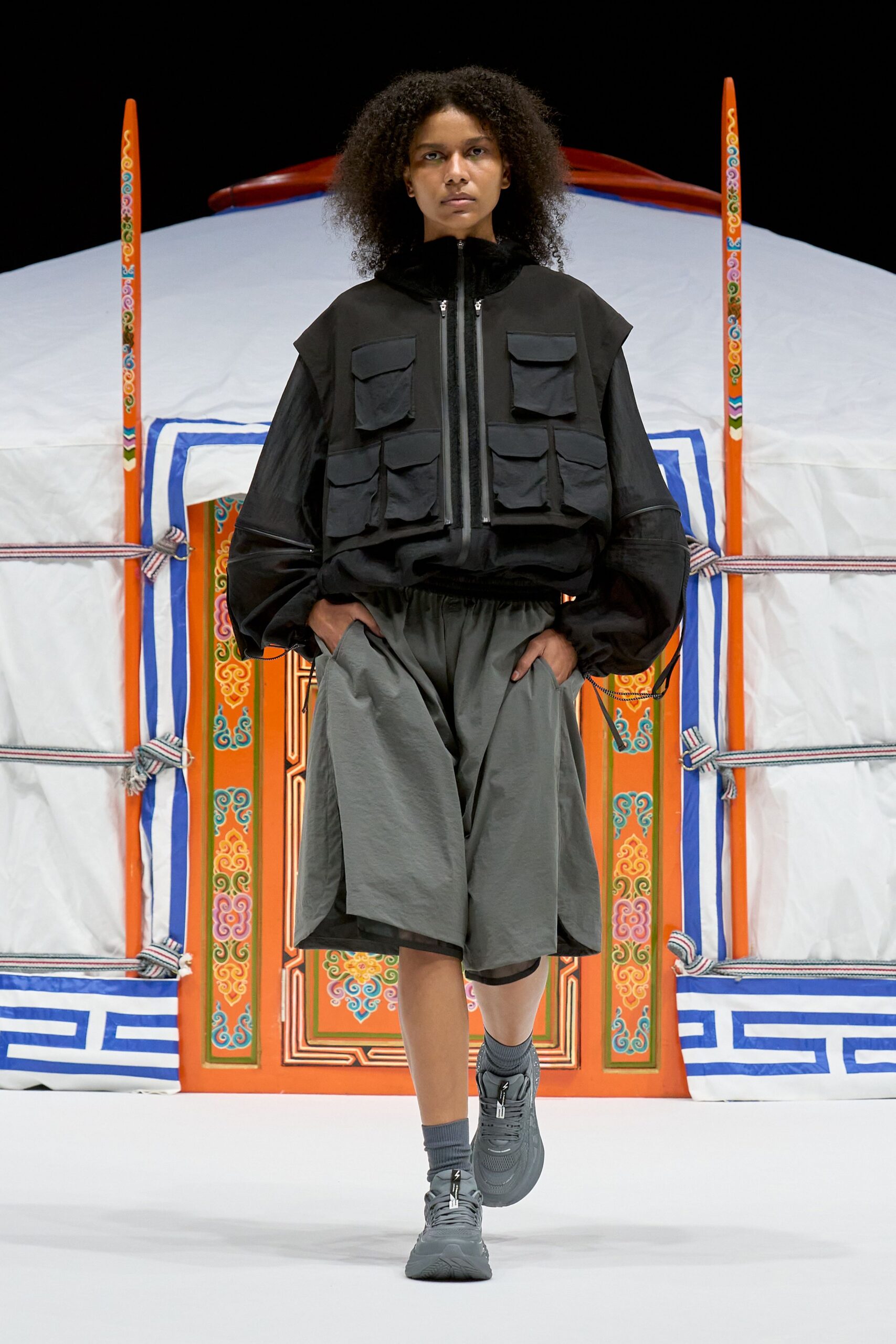 Black utility vest with matte flap pockets over a black windbreaker, its sleeve zips opening at the elbows. Charcoal nylon culottes with a gathered waist, ribbed socks, grey trainers. Outside a yurt before sunrise, air thin enough to sting.