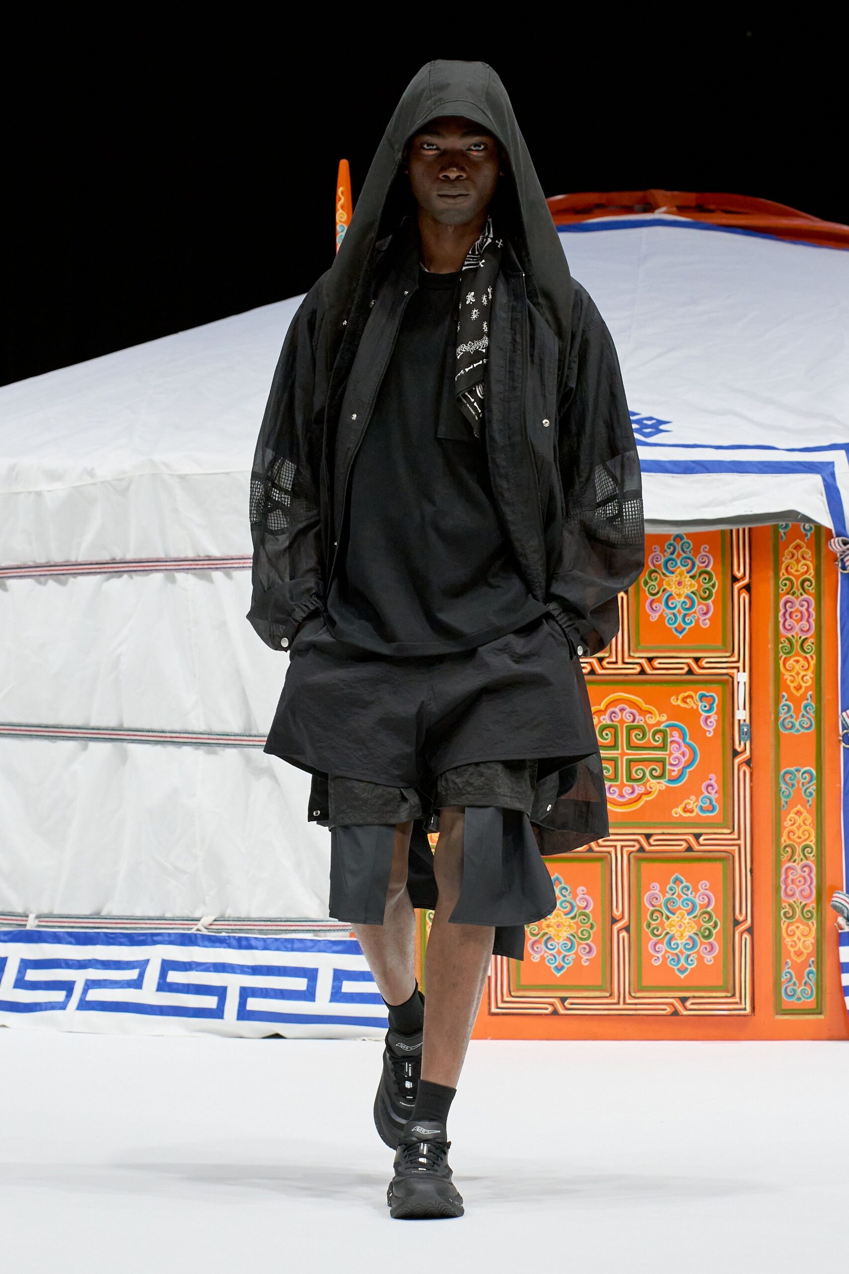 Black hooded parka with mesh insets at the sleeves, a black bandana-print scarf, loose black tee, paper-nylon overshorts over longer raw-edged shorts, black trainers. Like stepping out of a tent at dawn before the stove catches.