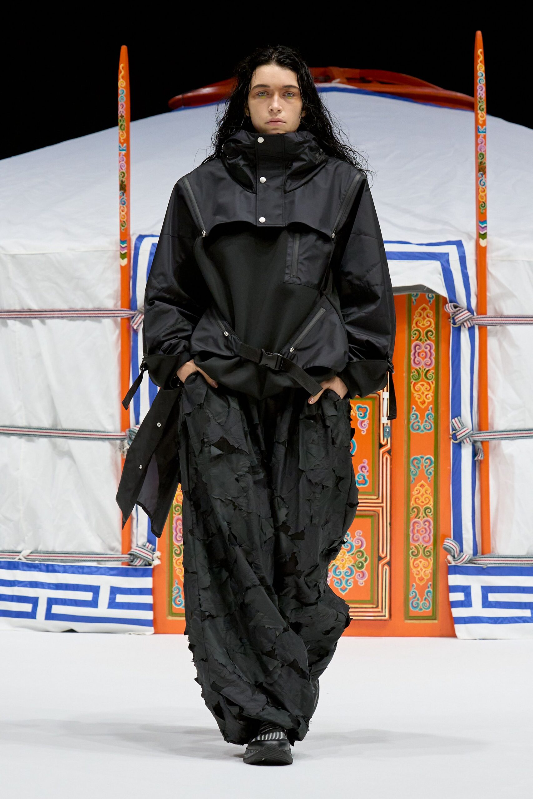 Black technical anorak with a snap-fastened storm flap and buckled waist strap, over a soot-dark skirt built from shredded appliqued layers. Ribbed black trainers peeking out below. Like stepping out to check the guy lines at dawn.